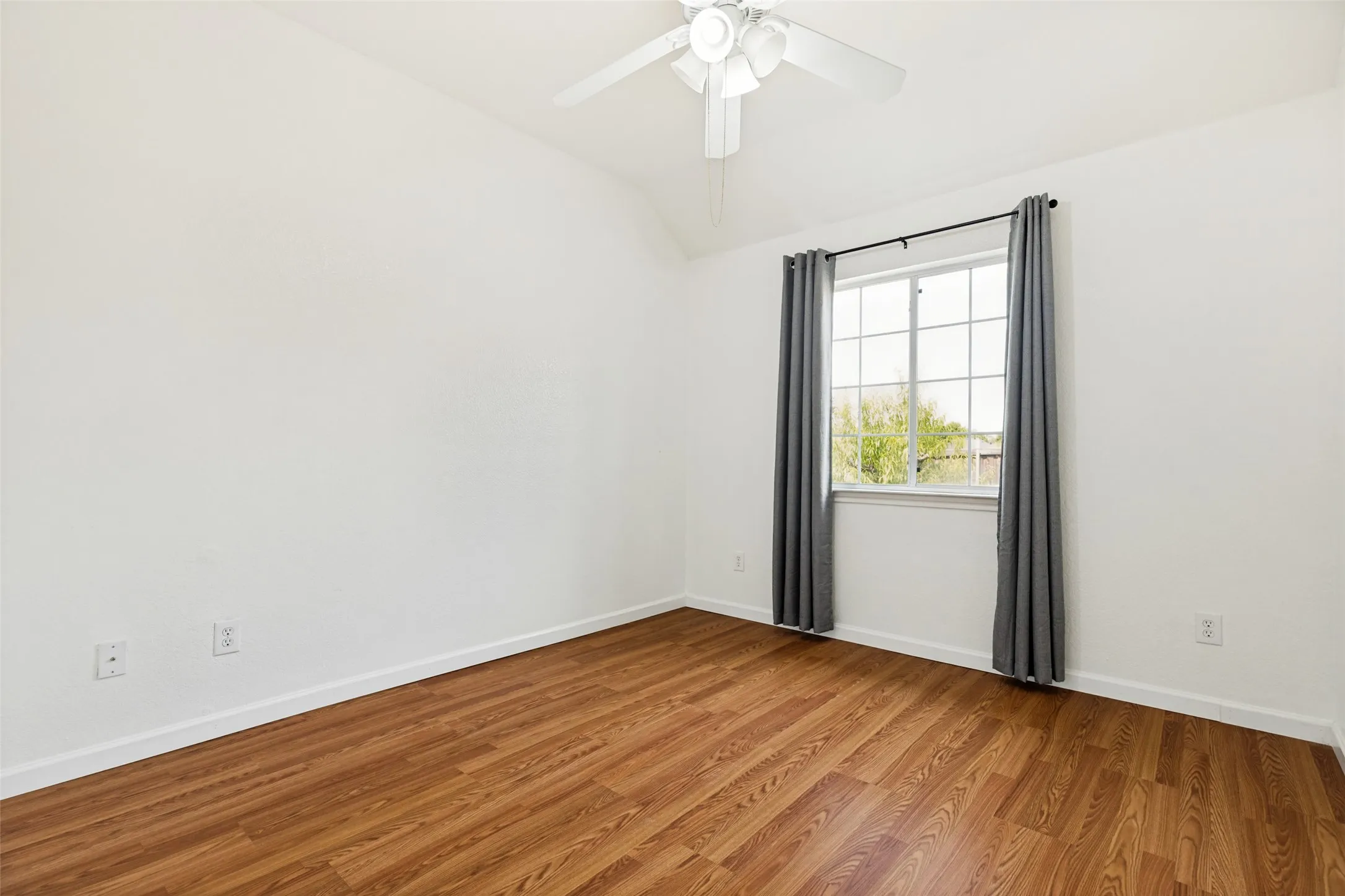 Vaulted ceiling, wood-finish flooring, white walls, ceiling fan, and a multi-pane window