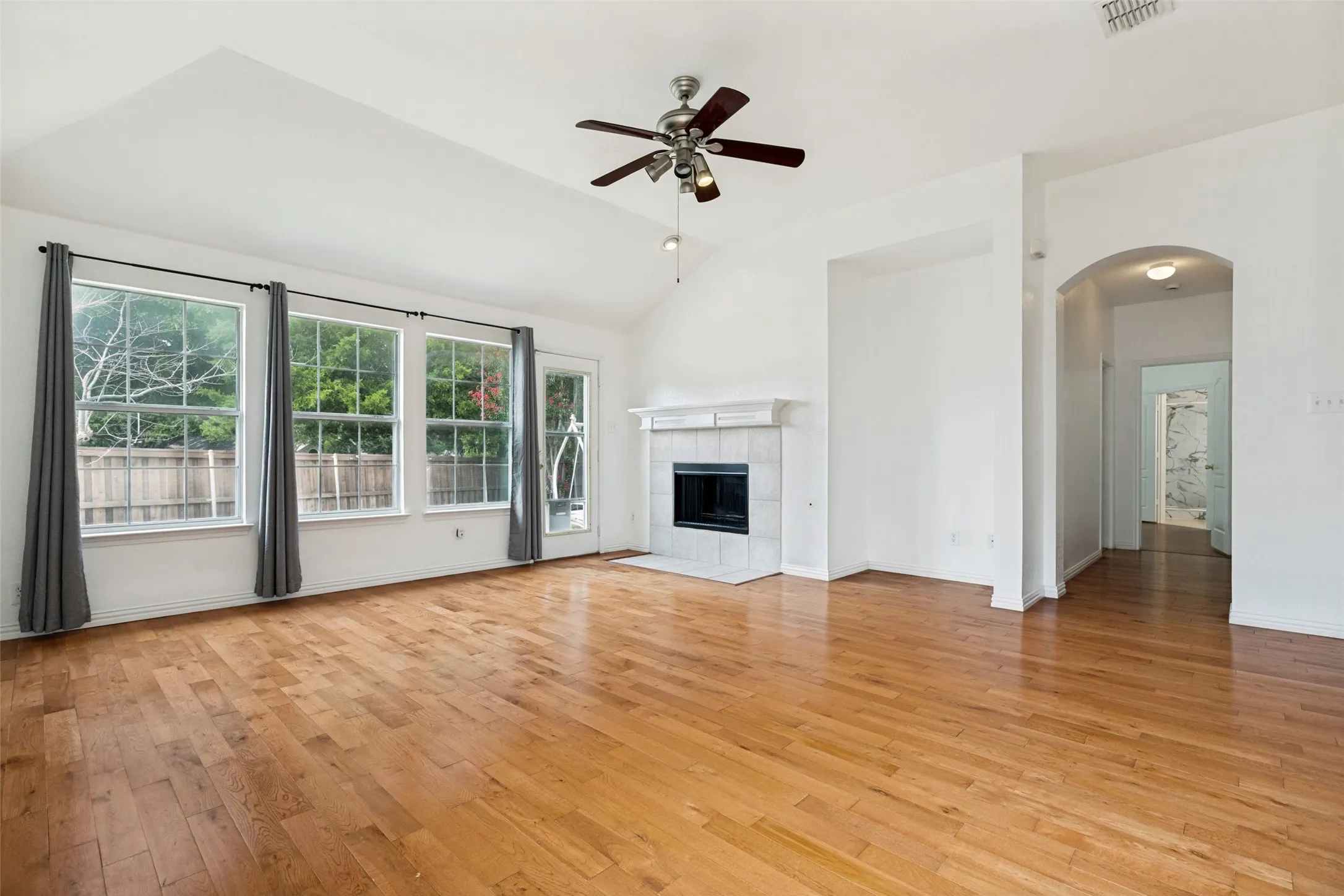 Vaulted ceiling with a ceiling fan, wood-finish flooring, a fireplace with a tile surround, multiple windows, and an arched doorway