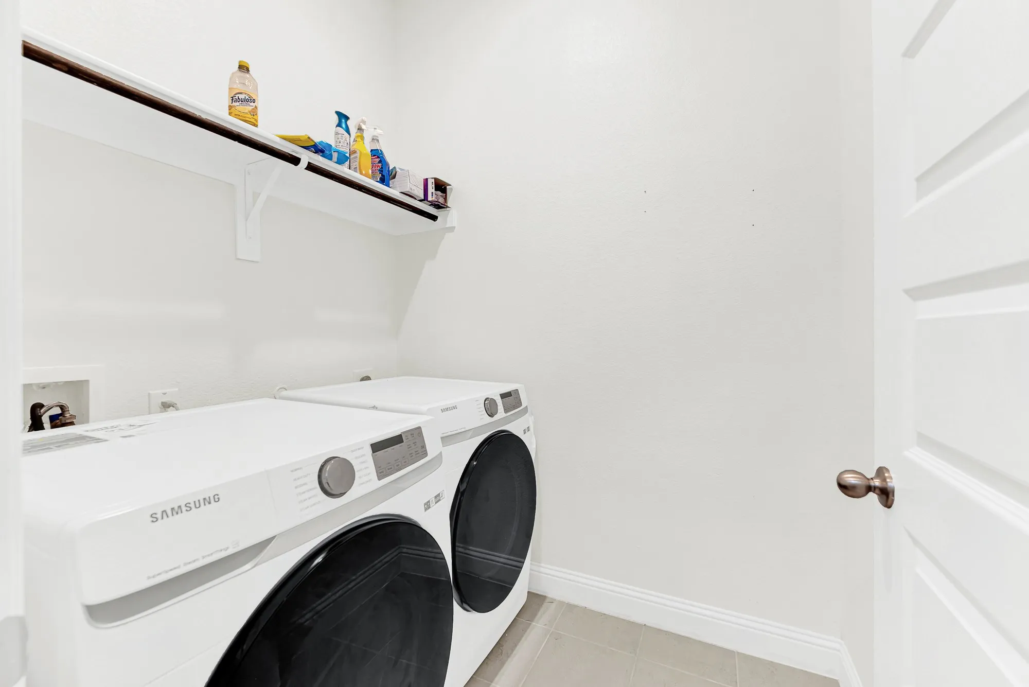 A dedicated utility space features high overhead shelving and bright neutral walls surrounding side-by-side front-loading laundry machines. Imagine breezes of warm, fresh linen making weekly household chores feel crisp, simple, and organized.