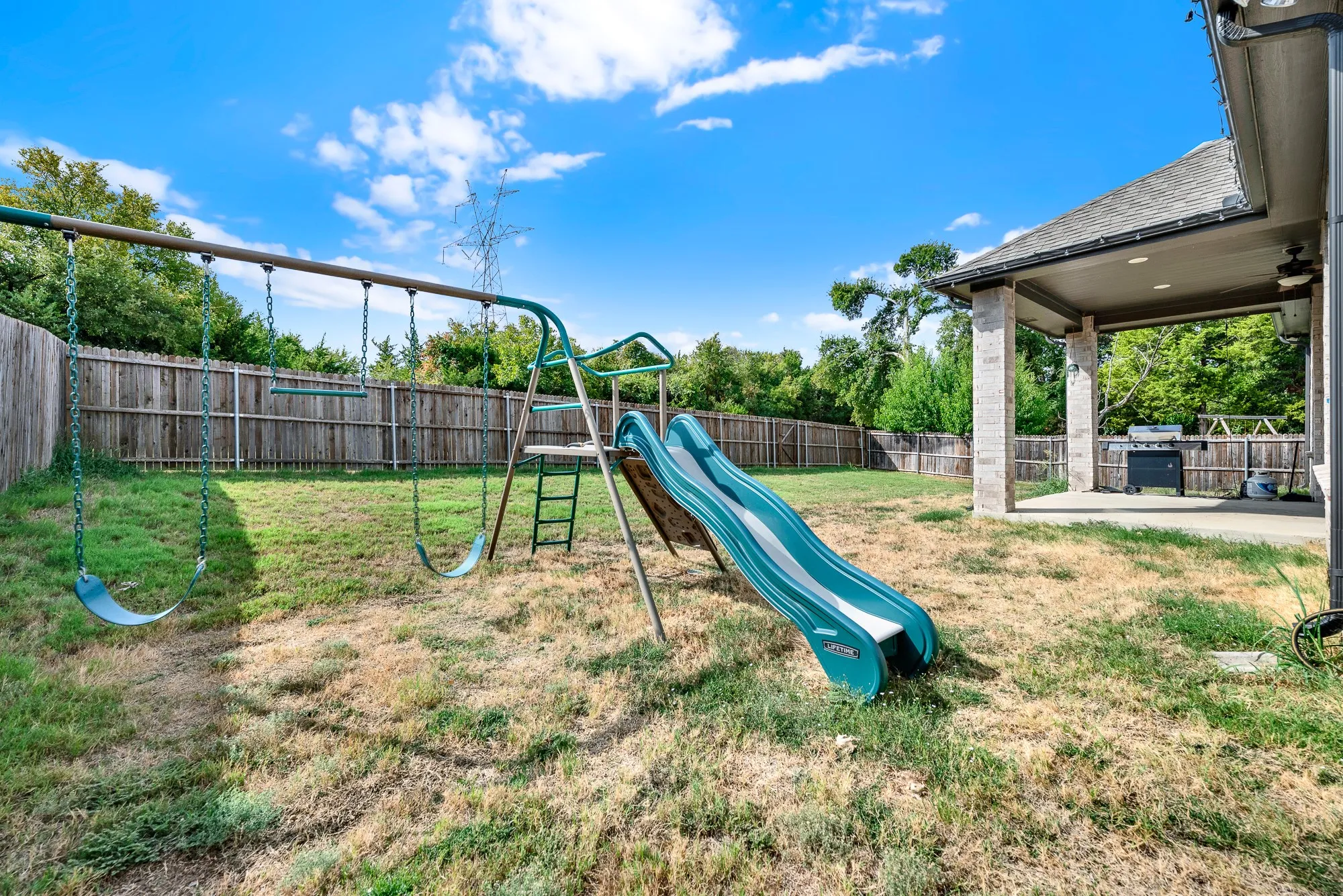 A tall covered patio with sturdy brick columns overlooks a deep, fully fenced lawn bordered by mature trees. Imagine watching kids play on the lawn while sipping cold drinks in the shaded outdoor shelter
