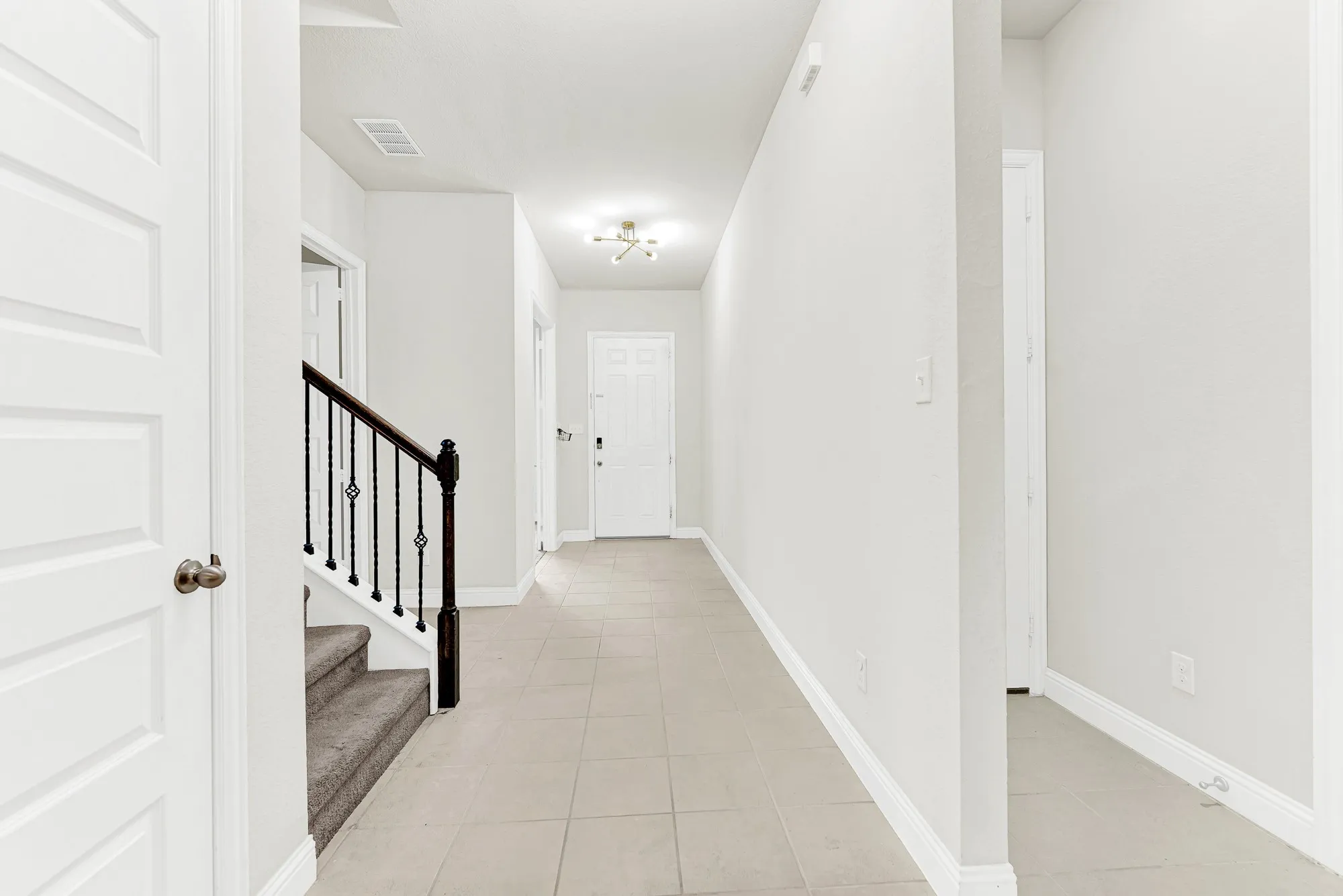 A long tiled hallway leads past a dark wood stair rail toward a modern sputnik light fixture that casts warm ambient light down the corridor. You can easily picture welcoming arriving guests with a warm greeting the moment they step through the front door.