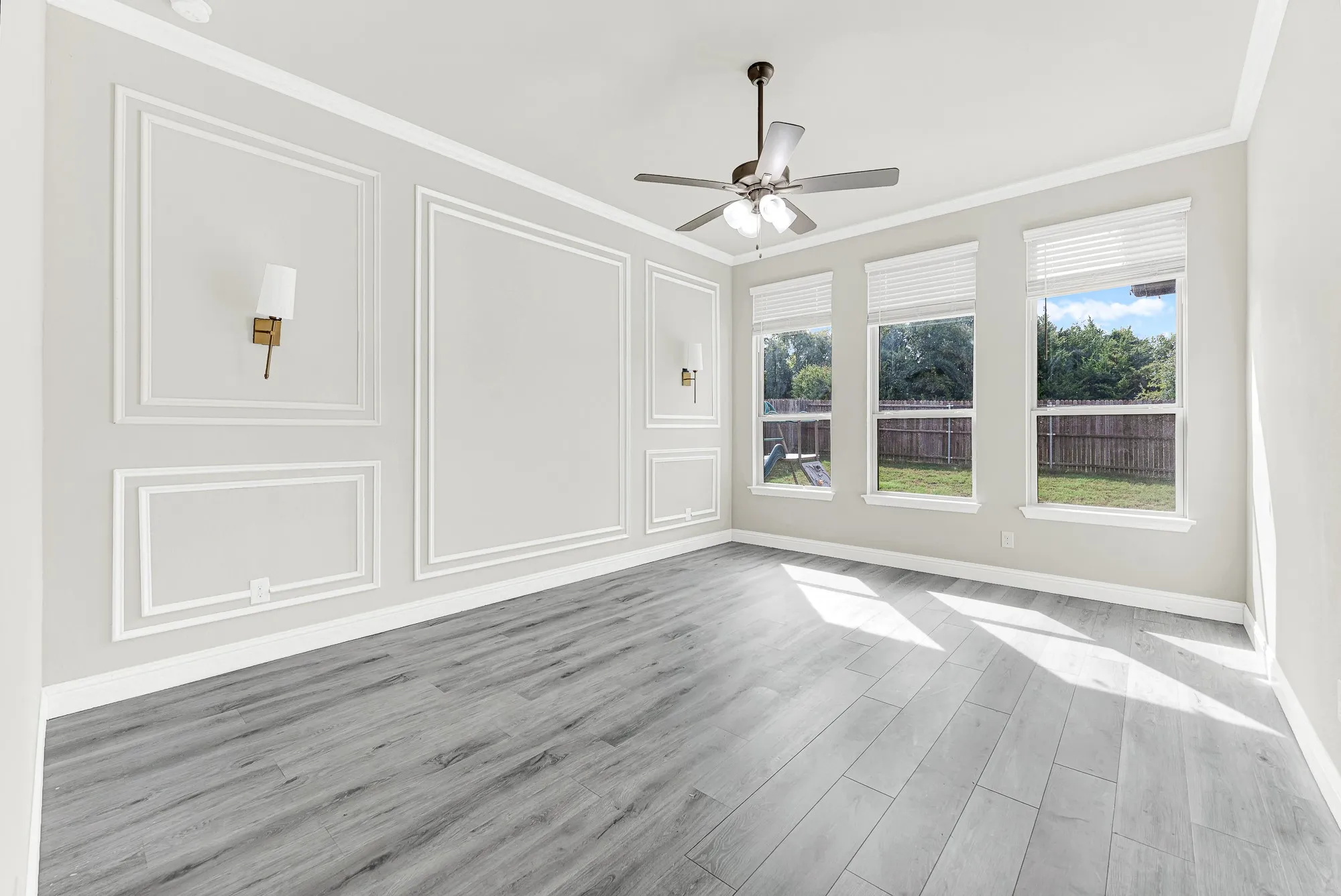 Elegant picture-frame wall molding and classic sconces flank a spacious focal wall opposite a trio of sunlit backyard windows. You can easily picture waking up to gentle morning sunbeams streaming across modern wood-look floors.