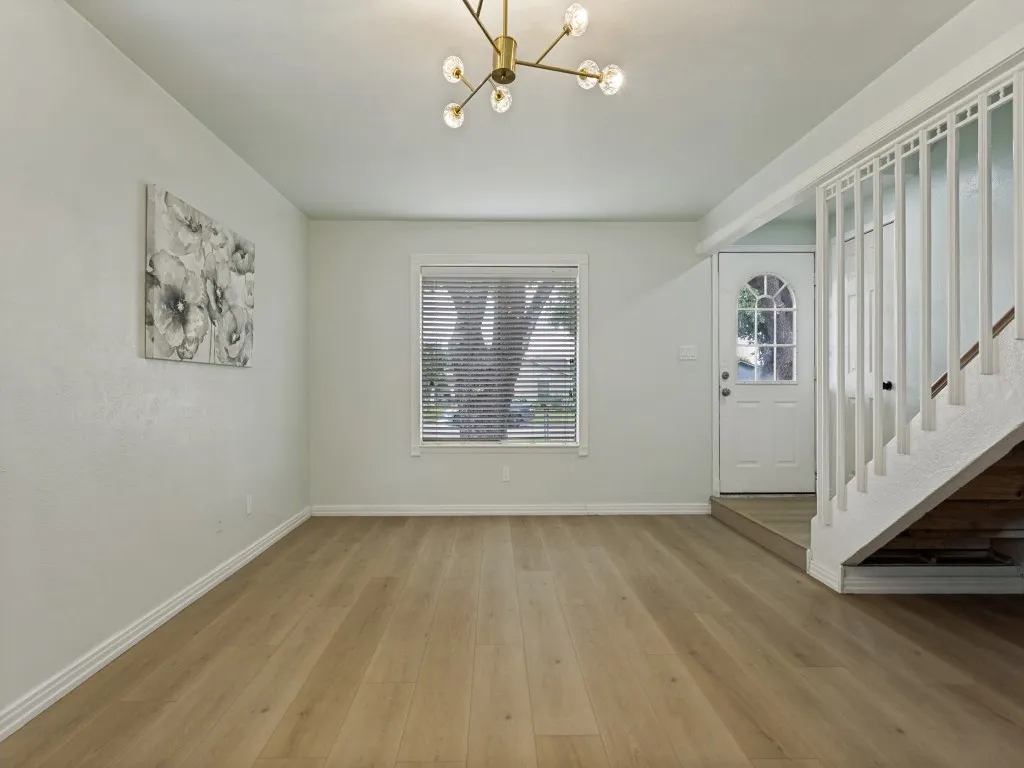 Wood-finish flooring throughout, modern chandelier, white staircase with vertical balusters, window with blinds, and an arched glass panel entry door
