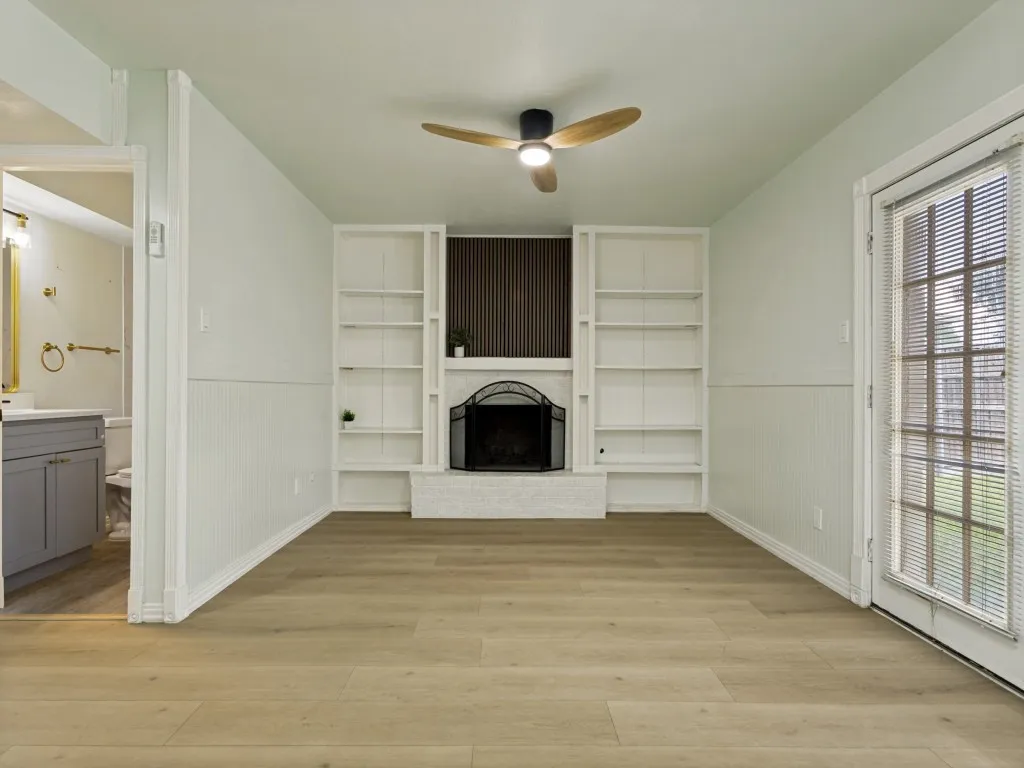 Wood-finish flooring, built-in shelving, a fireplace with a decorative screen, a ceiling fan with integrated lighting, and a glass-paned door