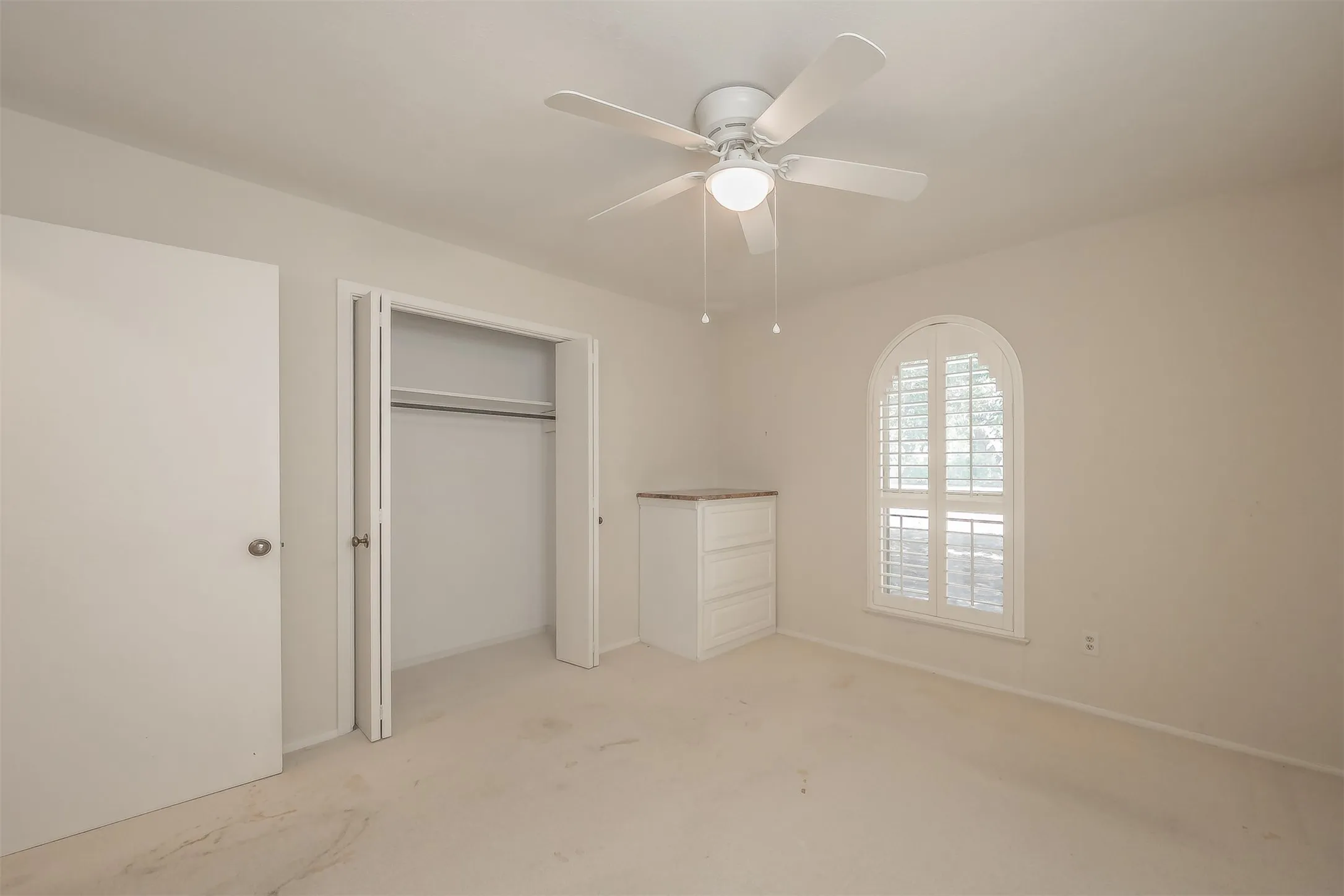 Arched window with plantation shutters, ceiling fan with integrated lighting, built-in dresser with a countertop, and a closet with bifold doors