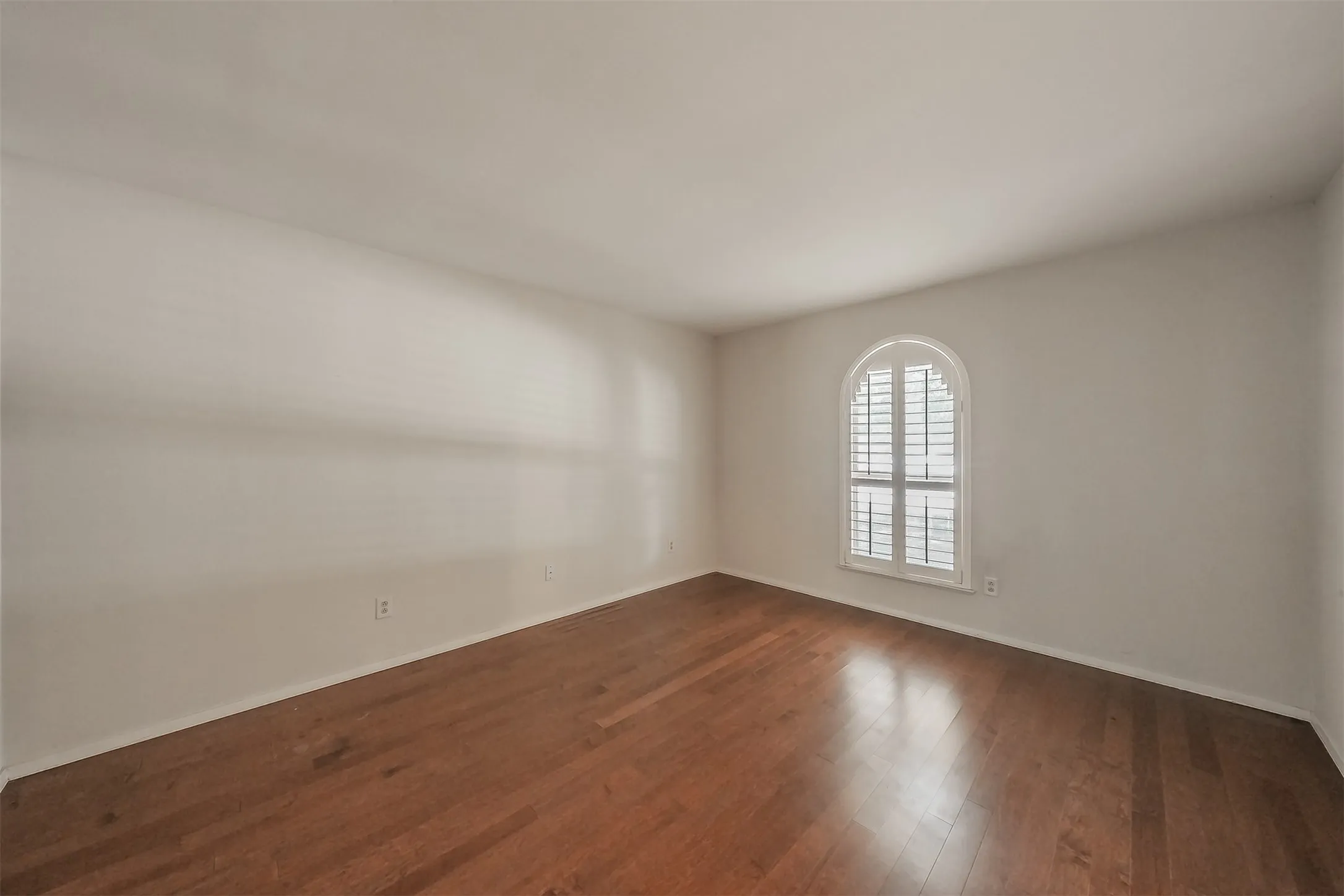 Wood-finish flooring, white walls, and a window with an arched top and louvered shutters