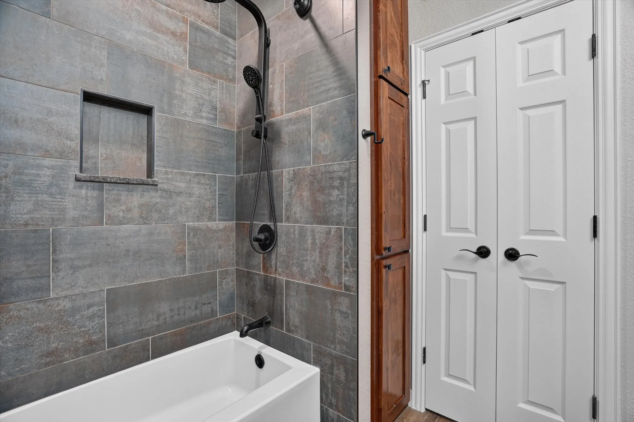 Bathtub with a matte black faucet, tiled shower, recessed shower niche, and a built-in wood-finish cabinet