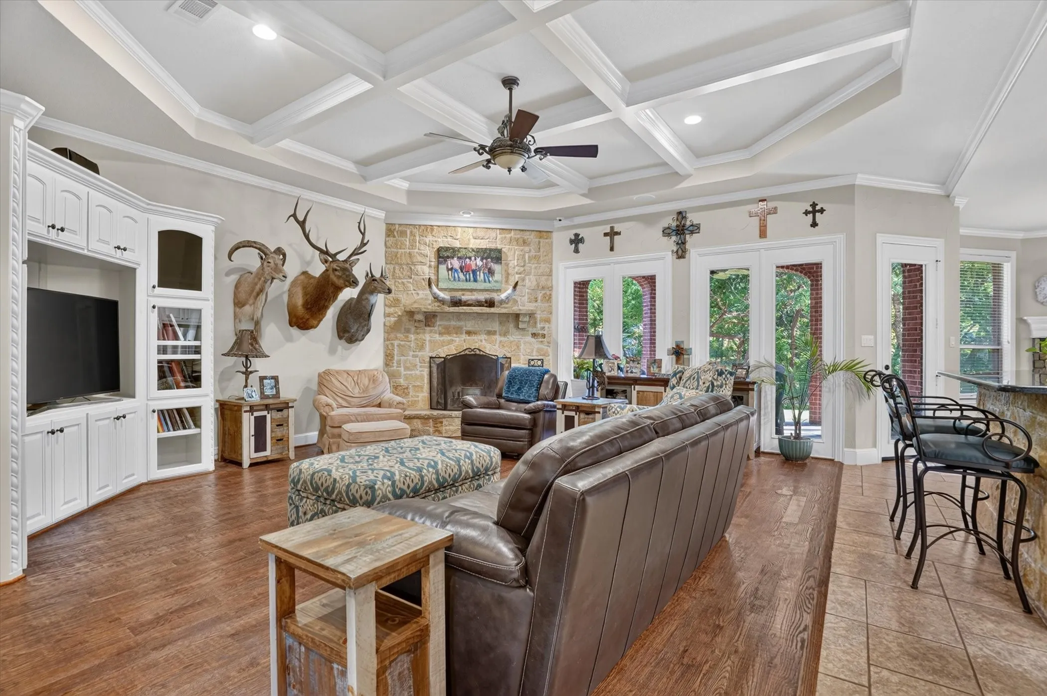 Coffered ceiling with a ceiling fan, stone-clad fireplace, hardwood flooring, built-in cabinetry, and multiple glass windows.