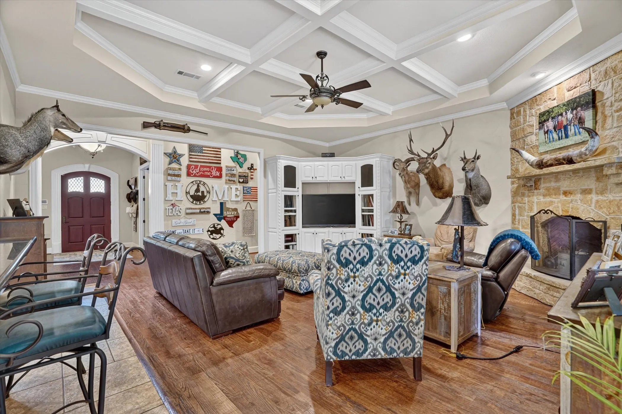 Coffered ceiling with recessed lighting and a ceiling fan, stone-clad fireplace, built-in media center with white cabinetry, hardwood-finish flooring, and arched entryways