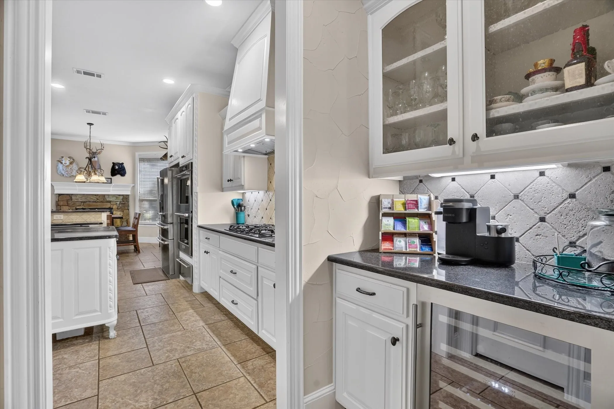 Ivory cabinetry with glass-front uppers, dark granite countertops, mini-fridge, and a patterned tile backsplash