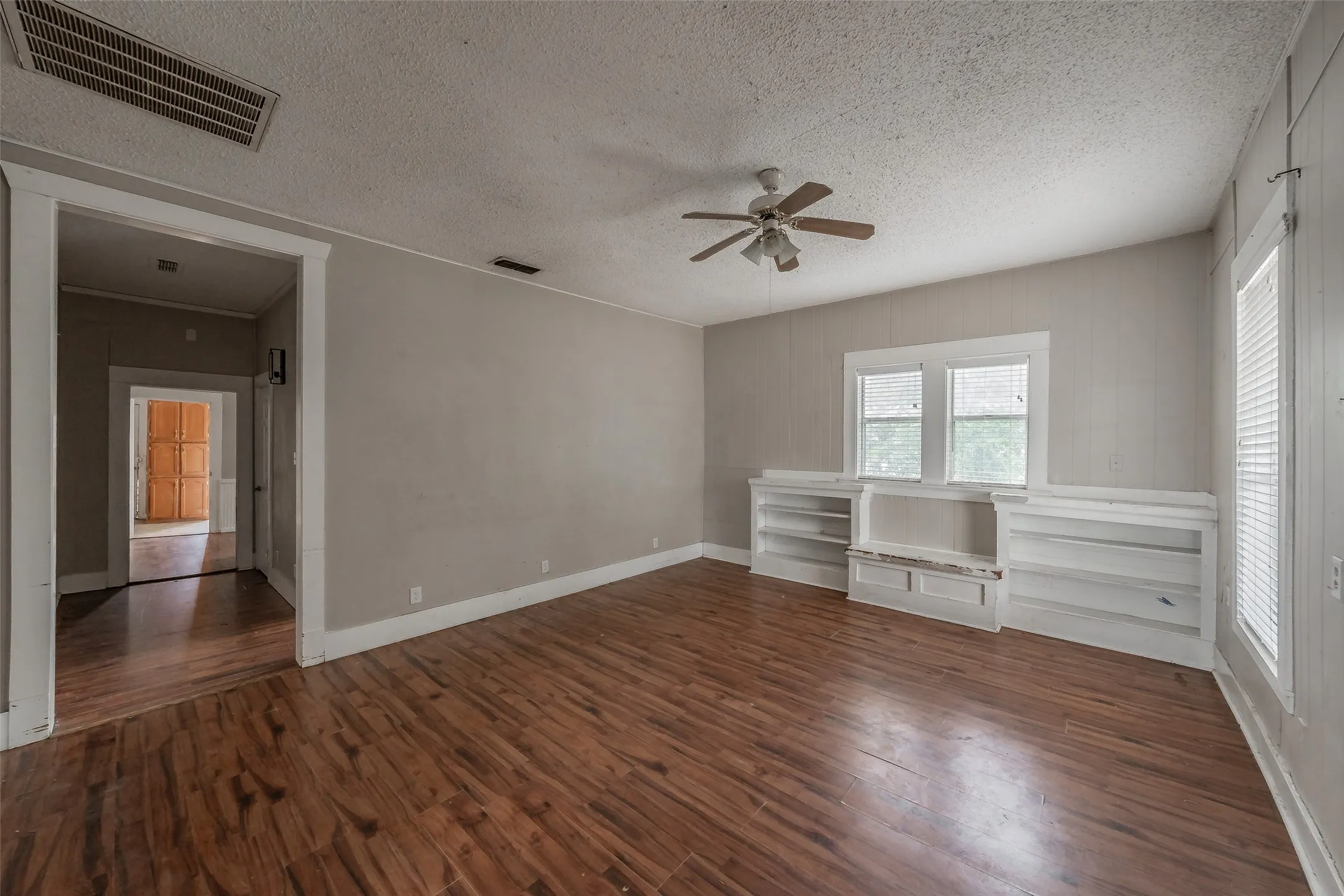 Wood-finish flooring, built-in shelving, two windows with white trim, and a ceiling fan with light fixture