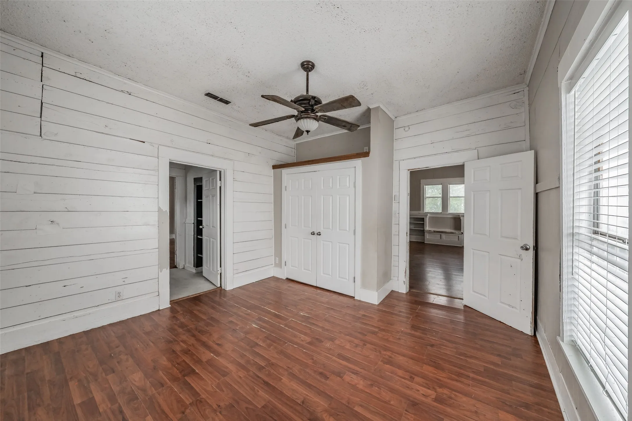 Wood-finish flooring, white shiplap walls, ceiling fan with light fixture, and white bi-fold closet doors