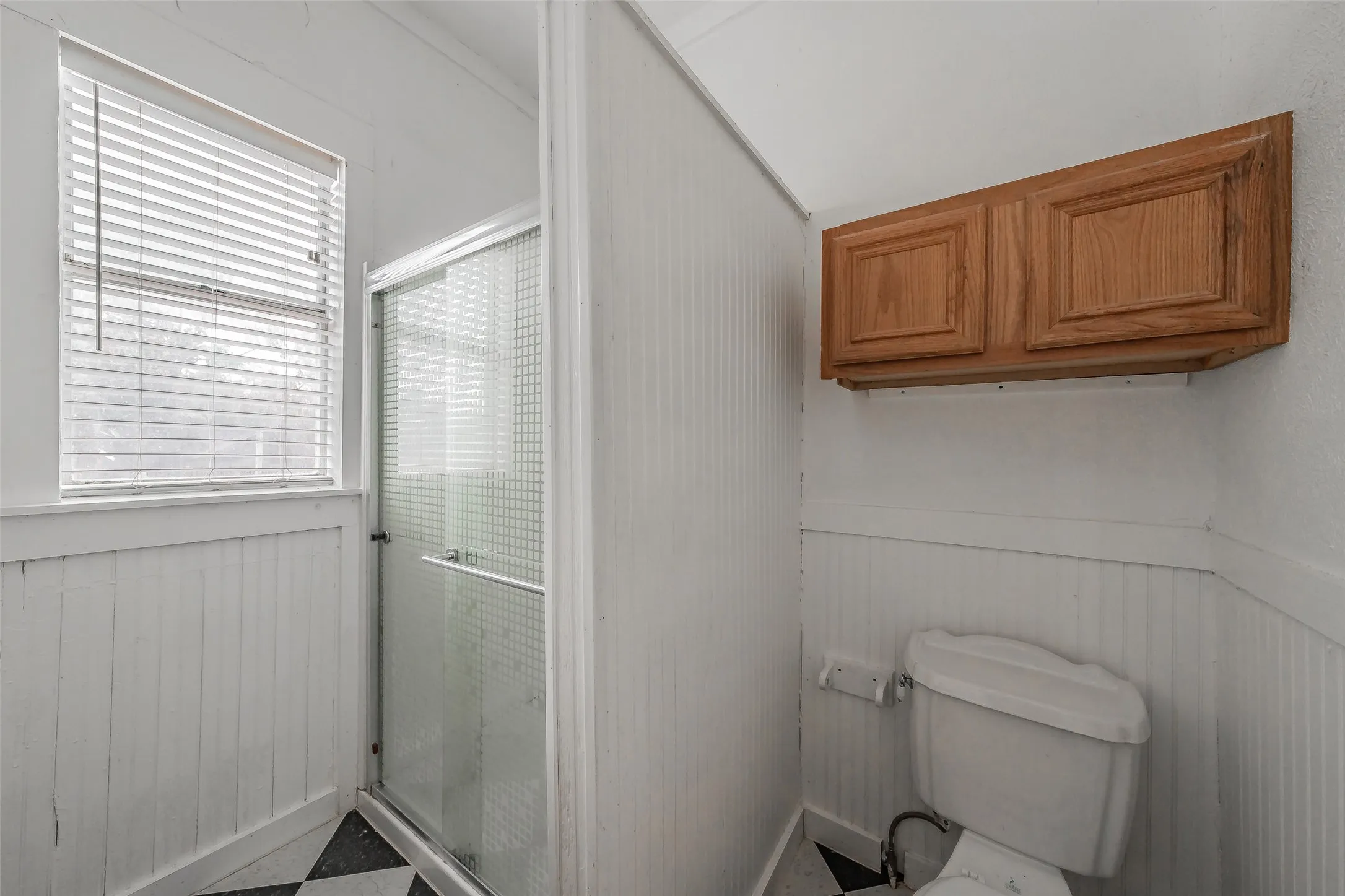 Shower enclosure with glass door, white beadboard paneling, black and white checkered flooring, and natural wood finish wall-mounted cabinetry