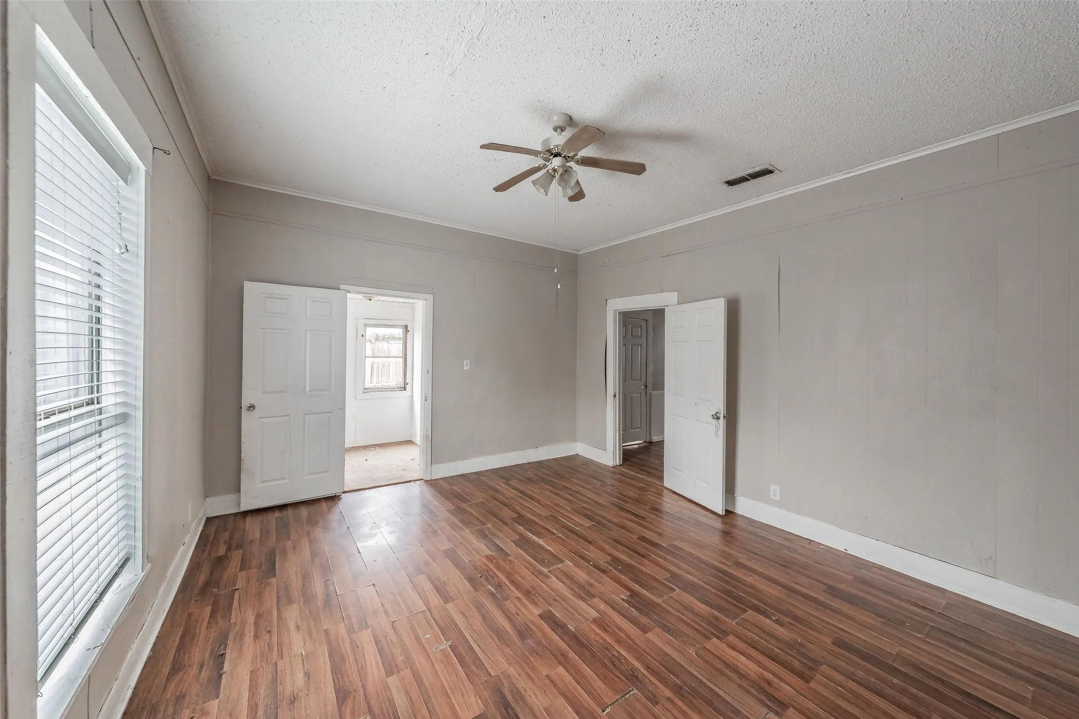 Wood-finish flooring, multiple paneled doors, window with blinds, and a ceiling fan