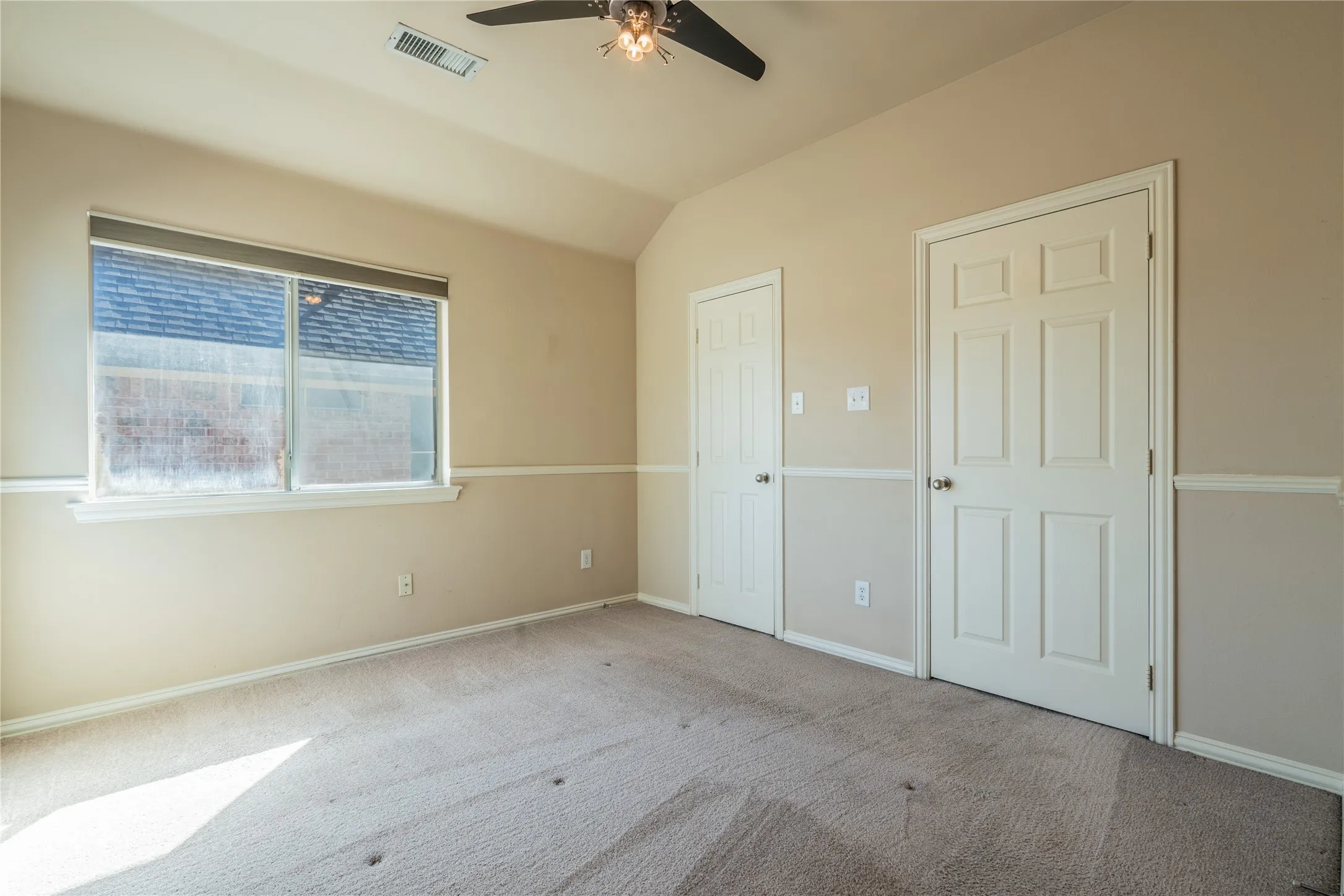 Another view of the bedroom with a vaulted ceiling with a ceiling fan, window with roller shades, two panel doors, and neutral carpeting