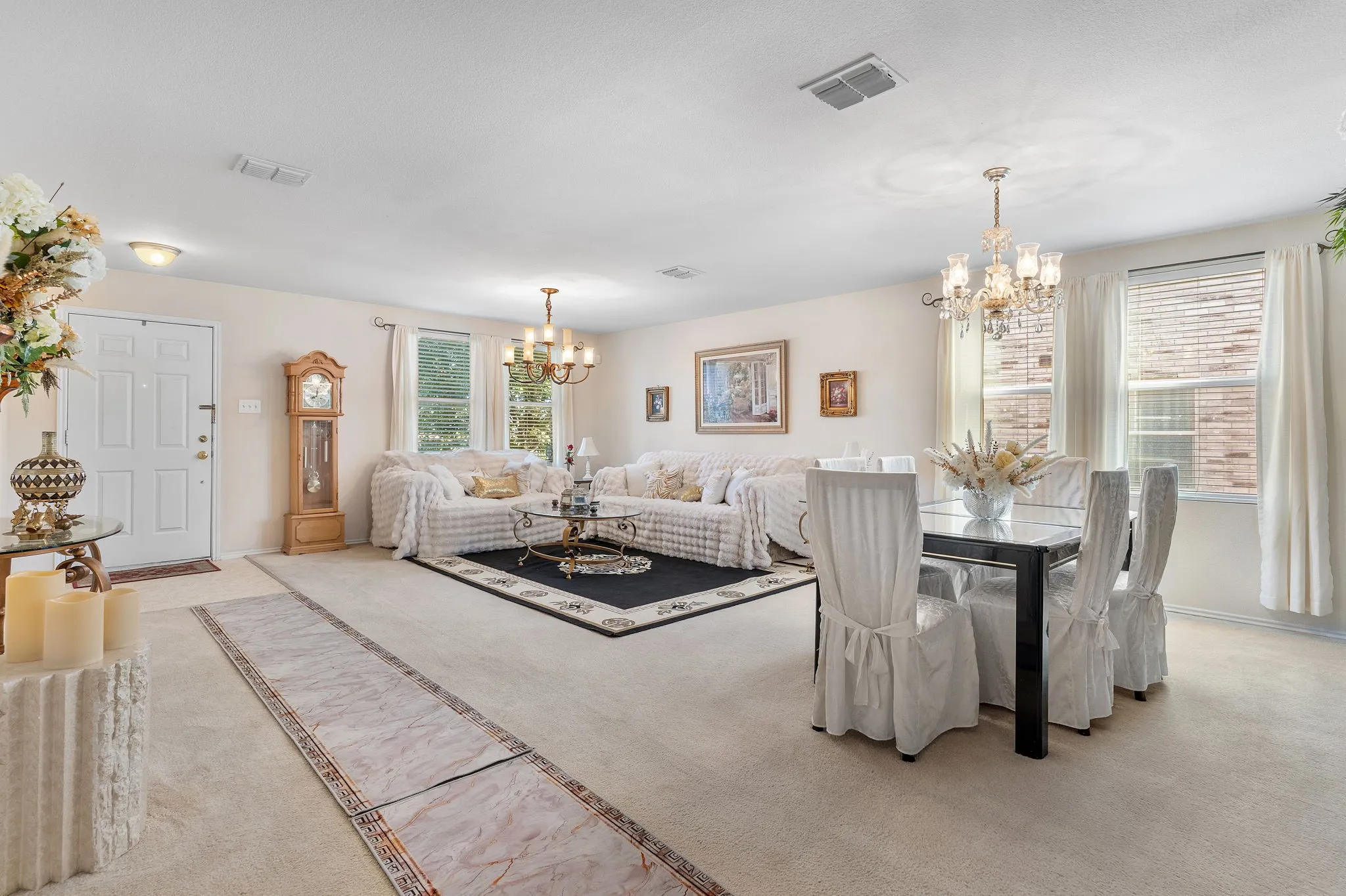 Open-concept living and dining area with neutral carpeting, multiple windows providing natural light, and two distinct chandelier light fixtures