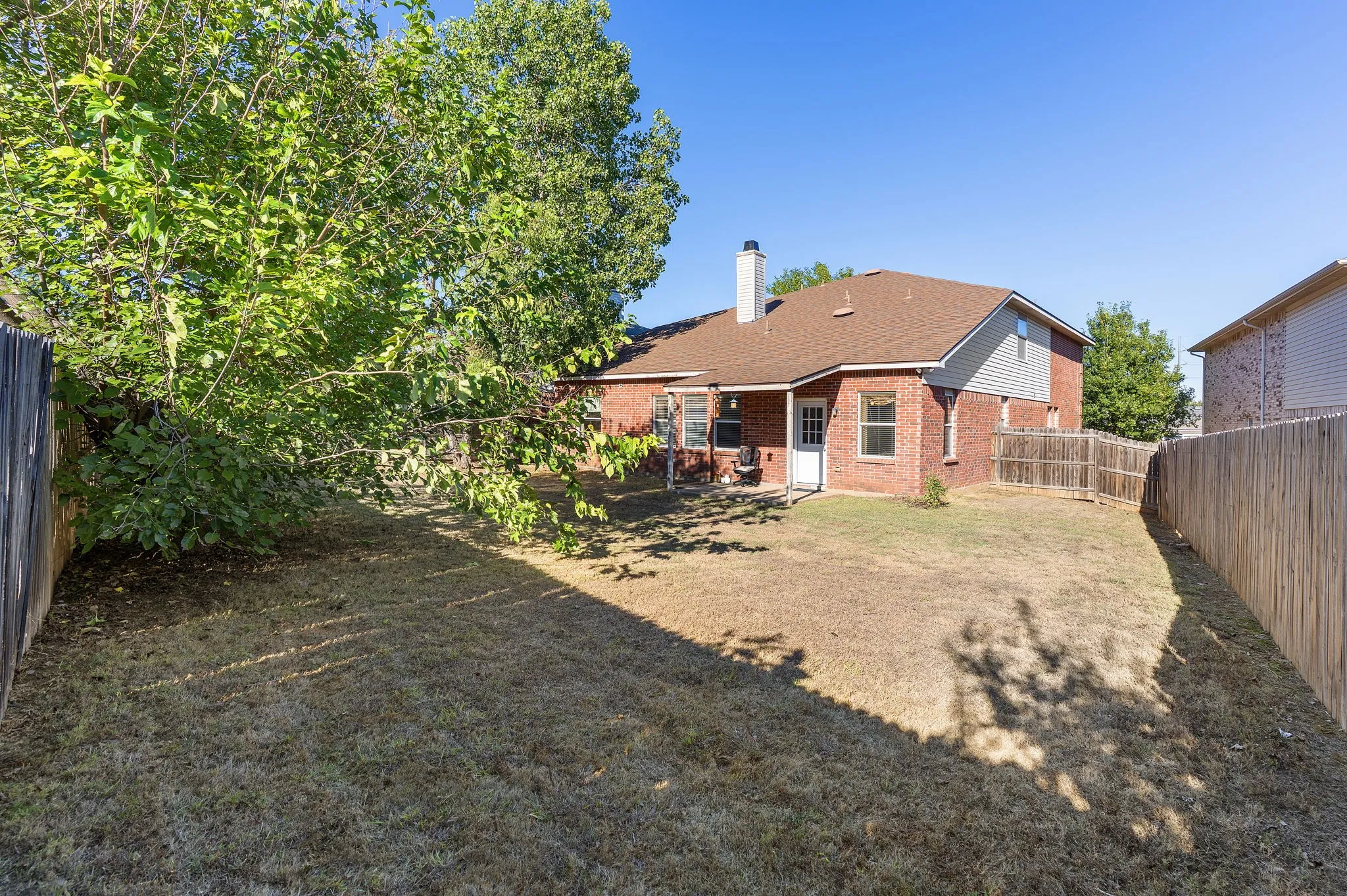 Expansive backyard featuring a privacy fence, mature trees, and a brick exterior with a white door