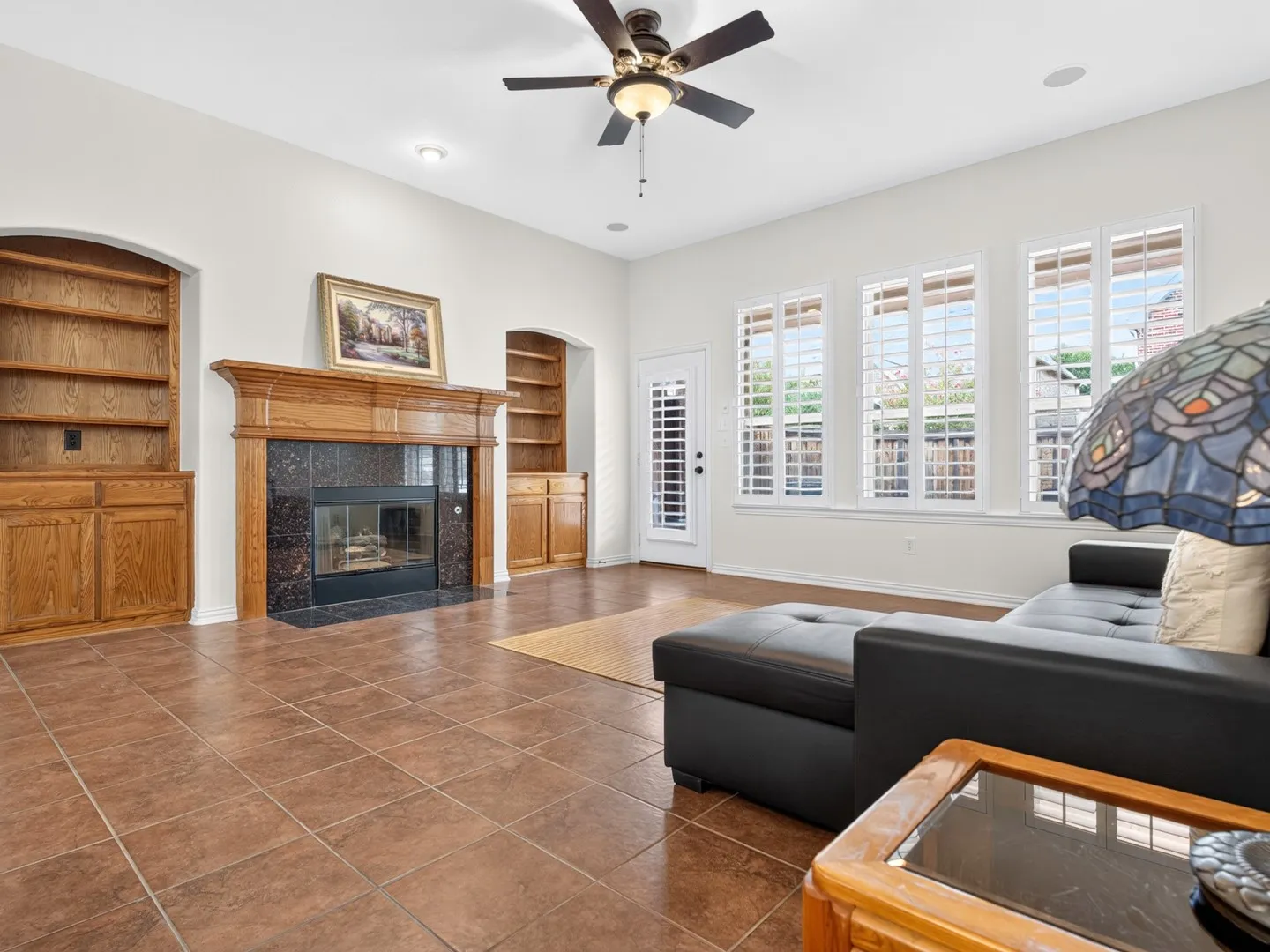 Spacious living area featuring tile flooring, a built-in fireplace with a stone surround, custom built-in shelving, multiple windows with plantation shutters, and a ceiling fan
