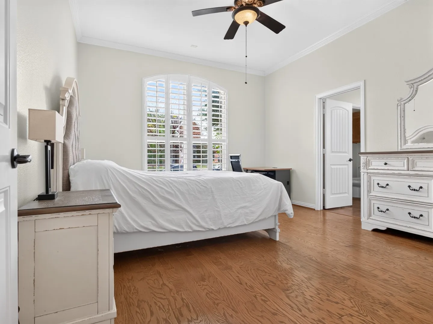 Wood-finish flooring throughout the room, arched window with plantation shutters, ceiling fan with integrated lighting, and crown molding
