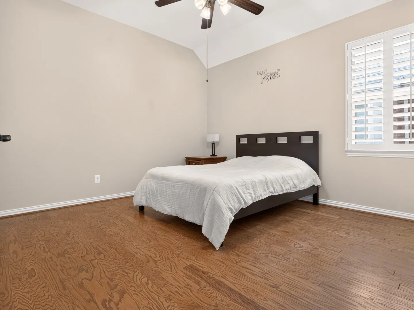 Vaulted ceiling with a ceiling fan, hardwood flooring, and window featuring plantation shutters