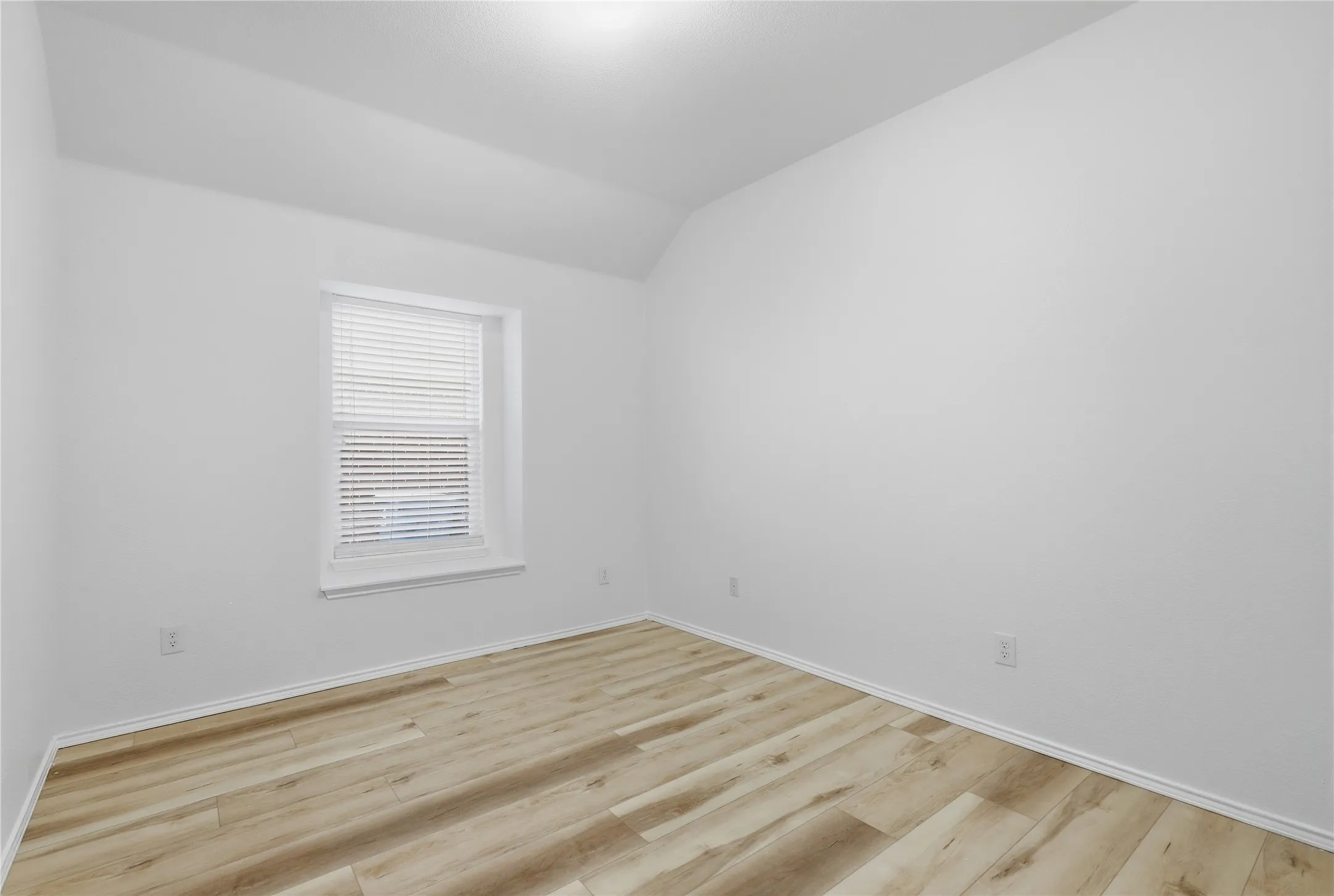 Wood-finish flooring with white trim, a single window with blinds, and white painted walls