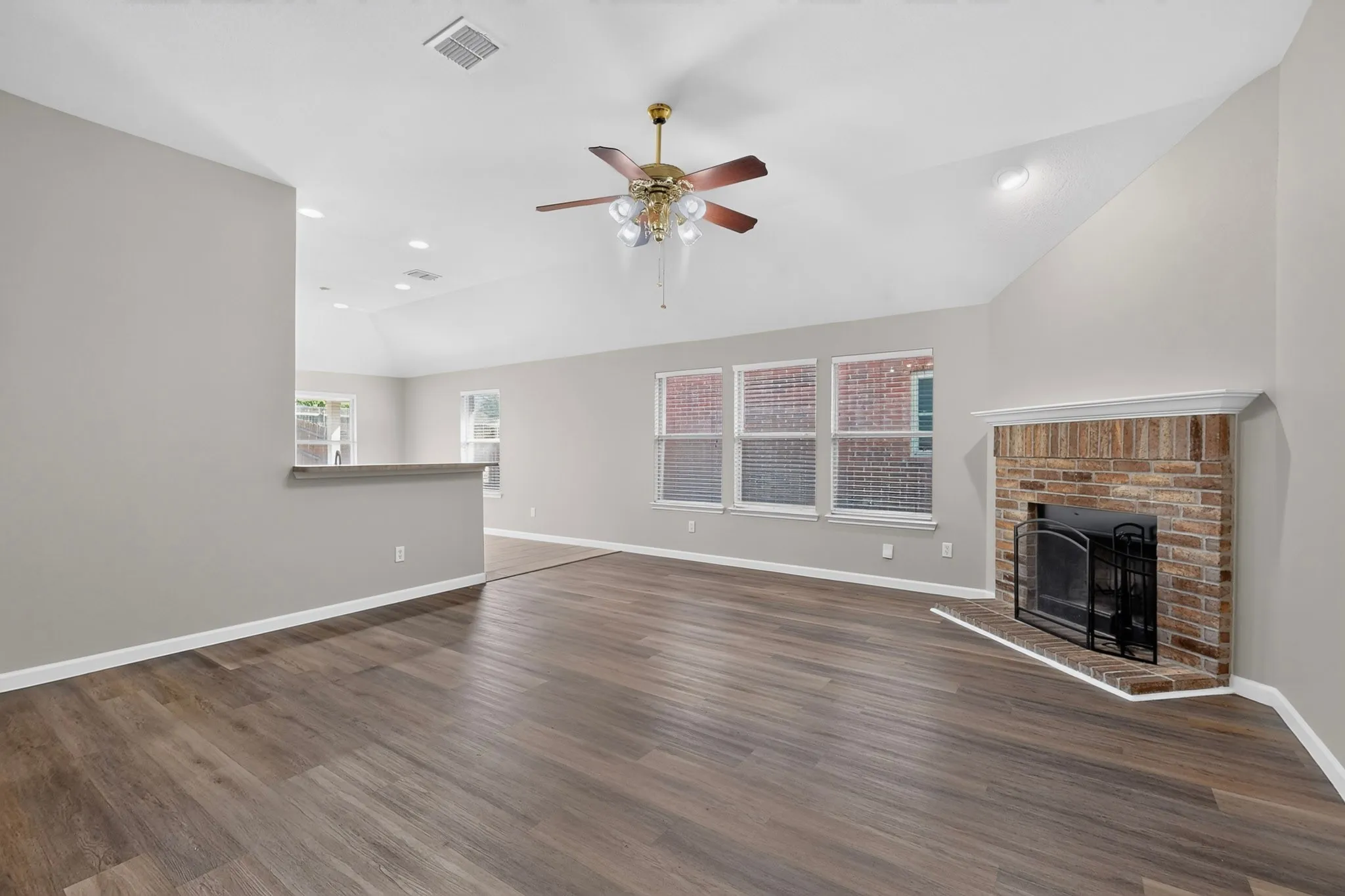 Vaulted ceilings featuring recessed lighting, wood-finish flooring, a brick fireplace with a white mantel, a ceiling fan with integrated lighting, and multiple windows