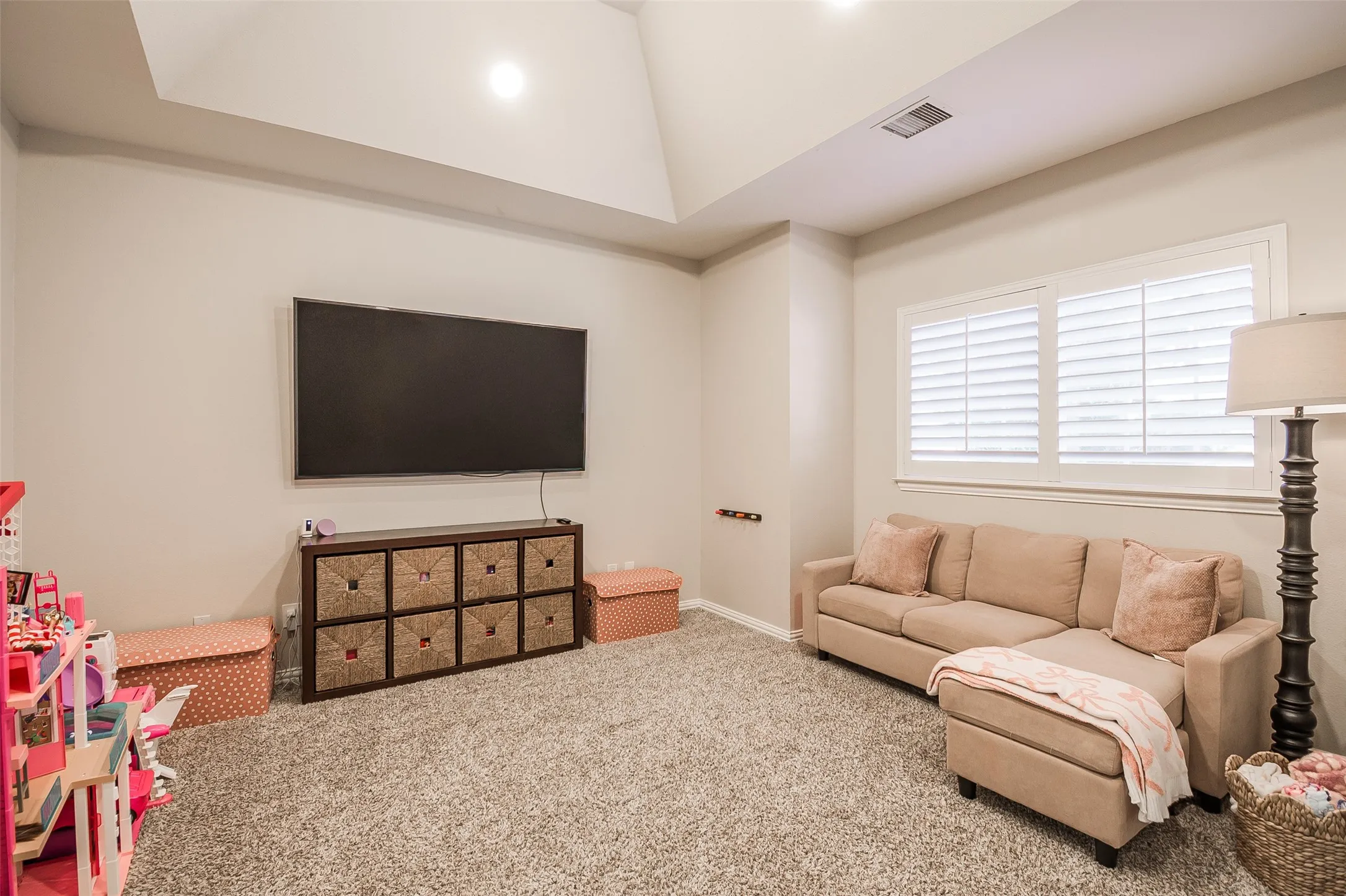 Carpeted room with a vaulted ceiling, wall-mounted television, and window with plantation shutters