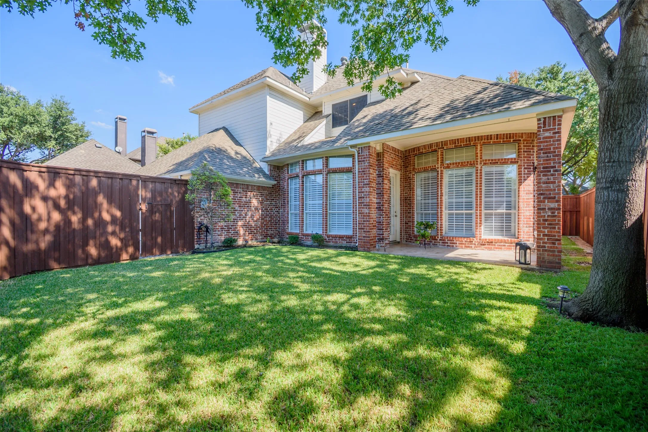 Expansive backyard featuring a manicured lawn, brick exterior, bay windows with shutters, and a covered patio