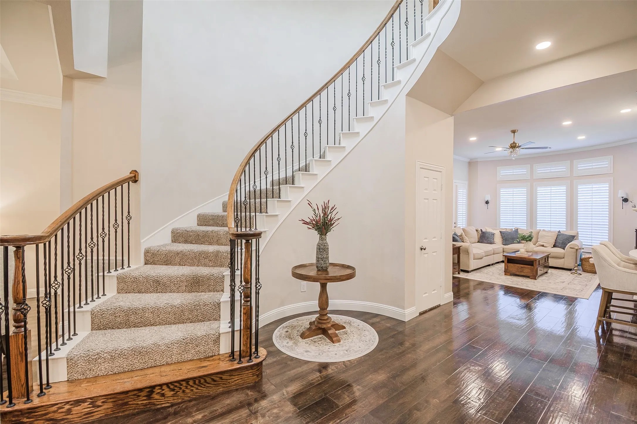 Sweeping staircase with wrought iron balusters and wood handrails, wood-finish flooring, recessed lighting, and a living area featuring window shutters