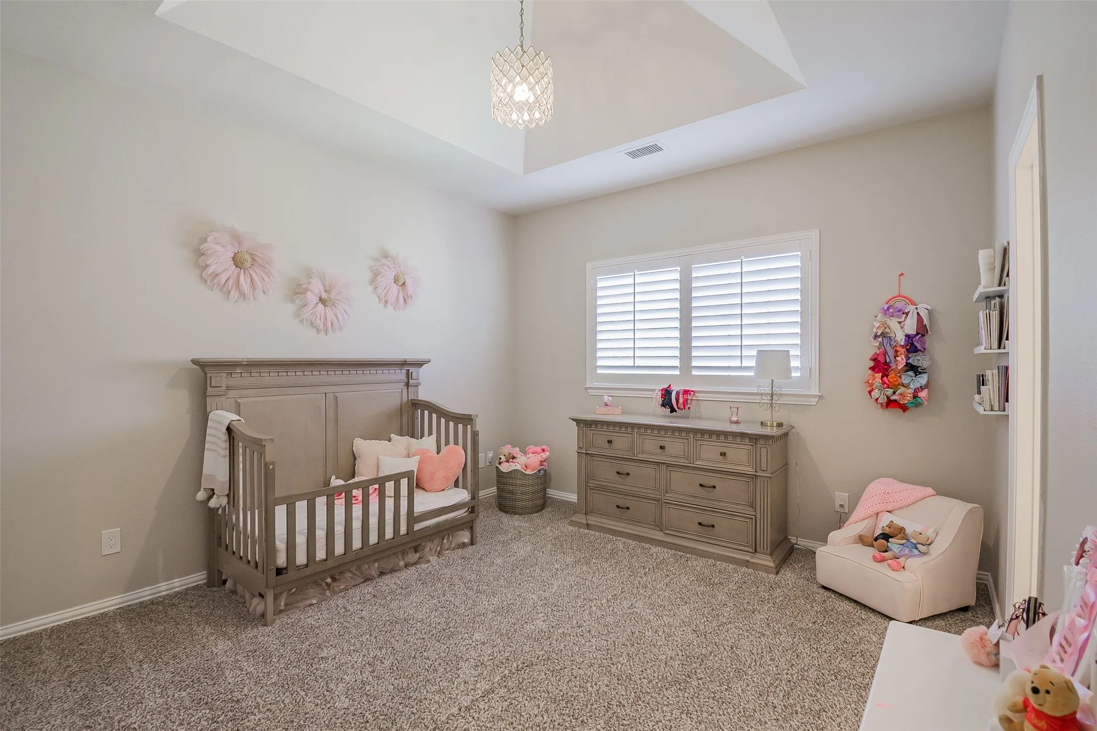 Bright nursery featuring a coffered ceiling with recessed lighting, plantation shutters framing large windows, neutral carpeting, coordinating wood-finish dresser and crib, and soft ambient décor throughout the space