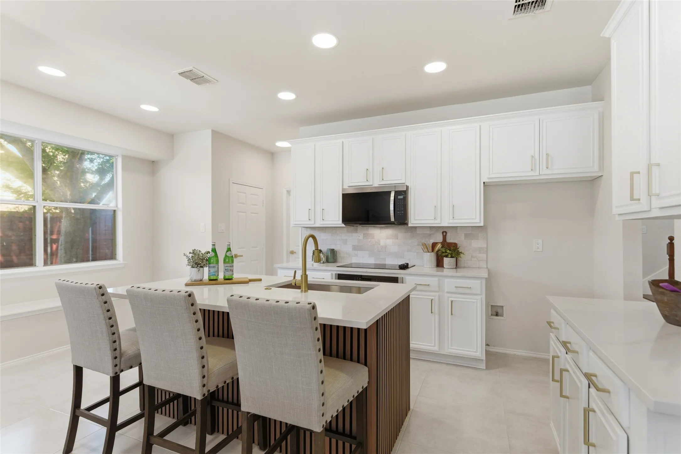 White cabinetry with gold-tone hardware, a central island with a built-in sink and gold-tone faucet, a white tile backsplash, and recessed ceiling lighting