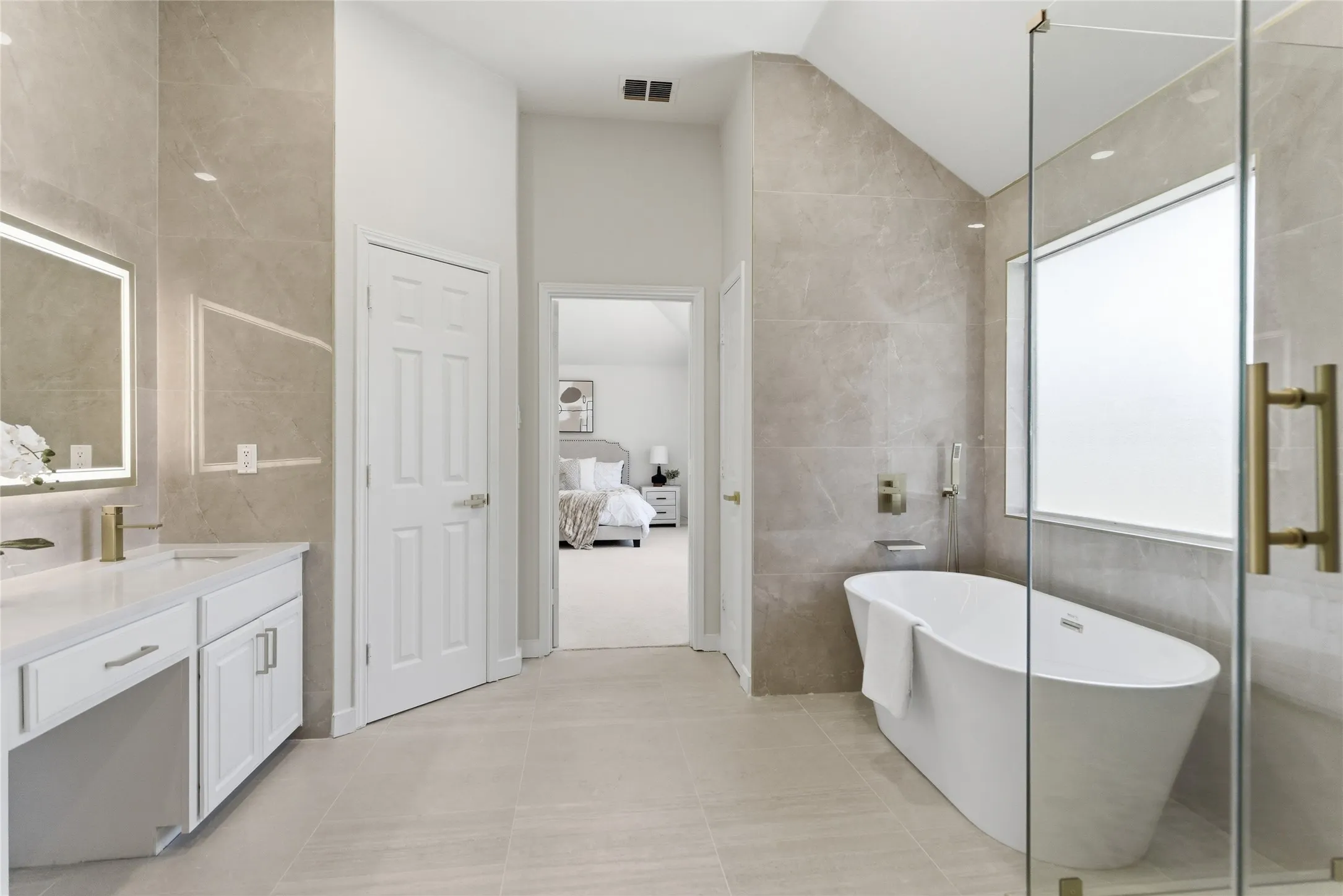 Freestanding soaking tub, frameless glass shower enclosure, vanity with integrated sink, illuminated mirror, and large format tile flooring