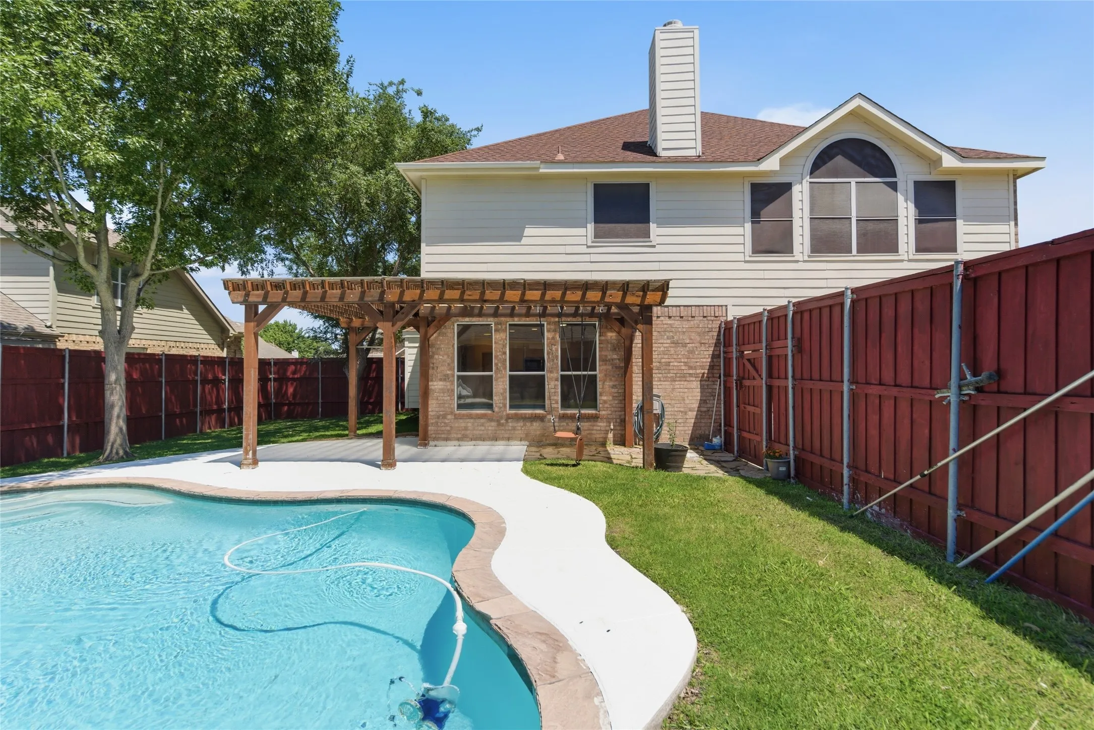 In-ground swimming pool with a surrounding concrete deck, wood pergola, and brick exterior accents