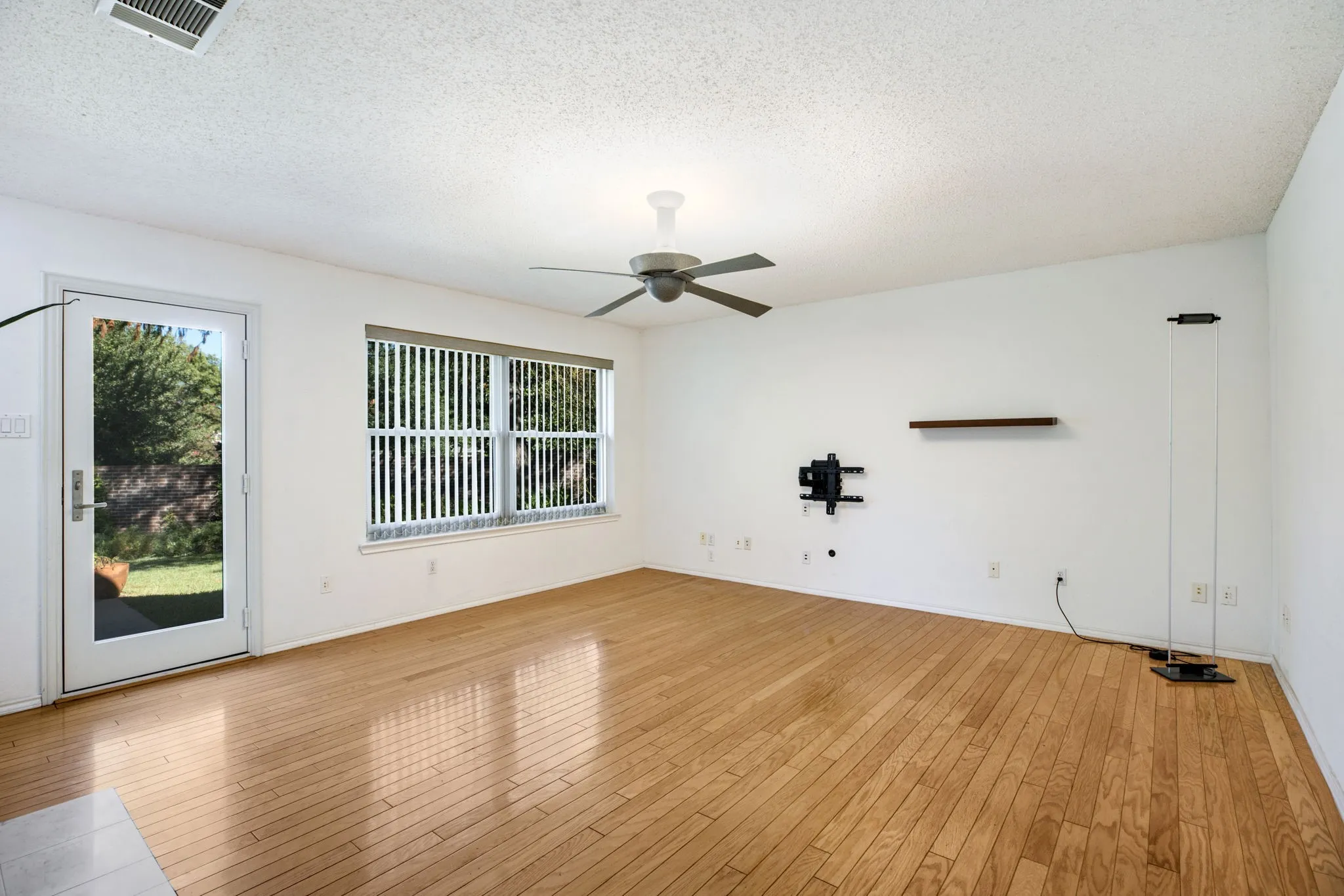 Wood-finish flooring throughout, ceiling fan, large window with vertical blinds, and a glass-paneled door