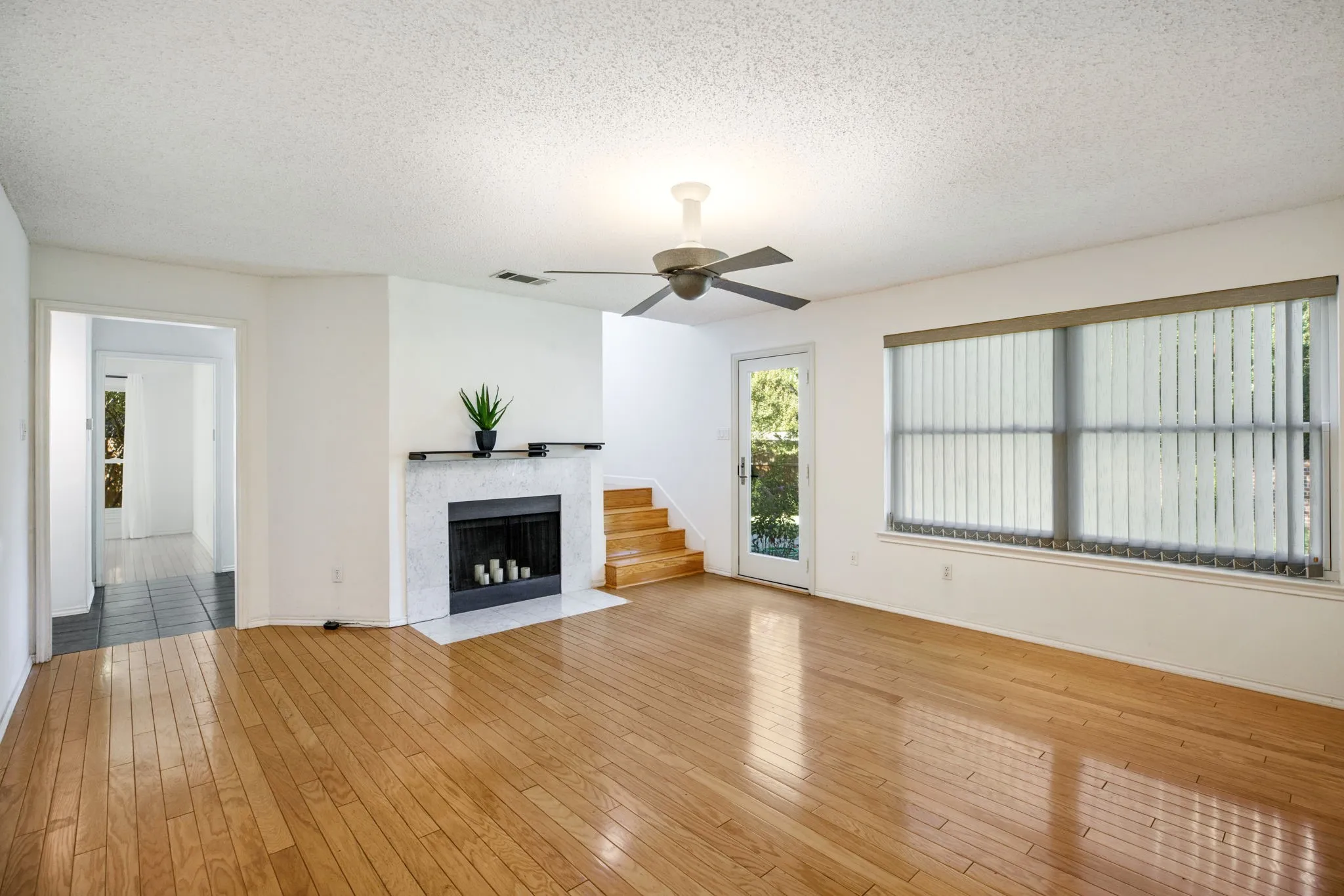 Light wood-finish flooring, a white-framed window with vertical blinds, a ceiling fan, and a fireplace with a white surround
