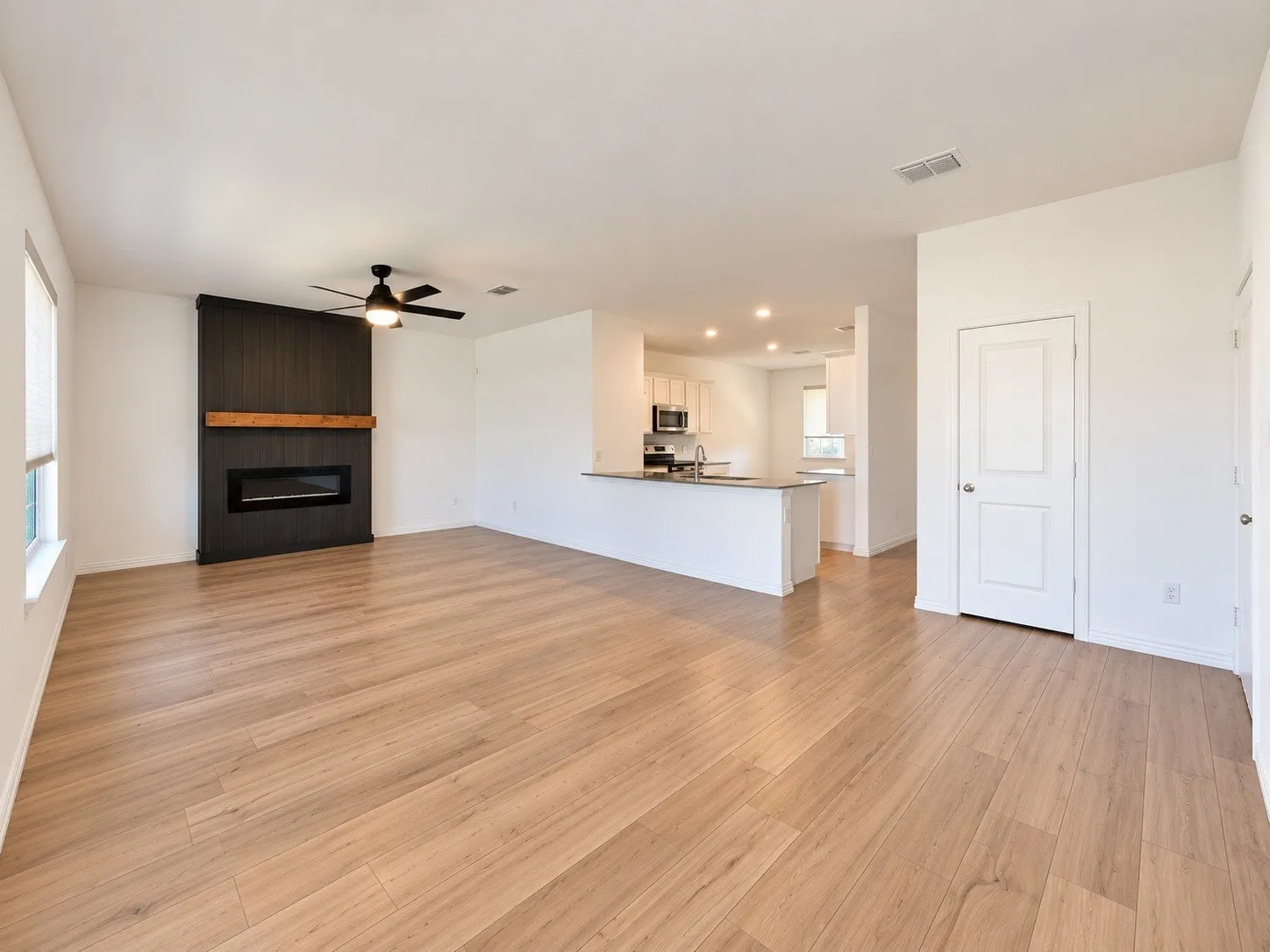 Spacious open-concept living area featuring wood-finish flooring, a modern fireplace with a dark wood-paneled surround and natural wood mantel, and a ceiling fan