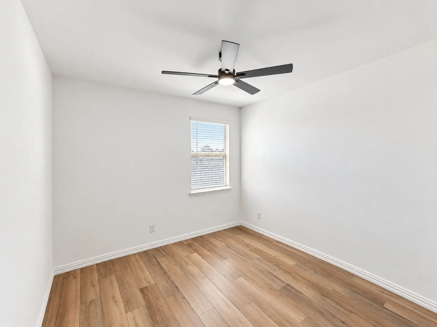 Wood-finish flooring with crisp white walls, a single window with blinds, and a ceiling fan with integrated lighting