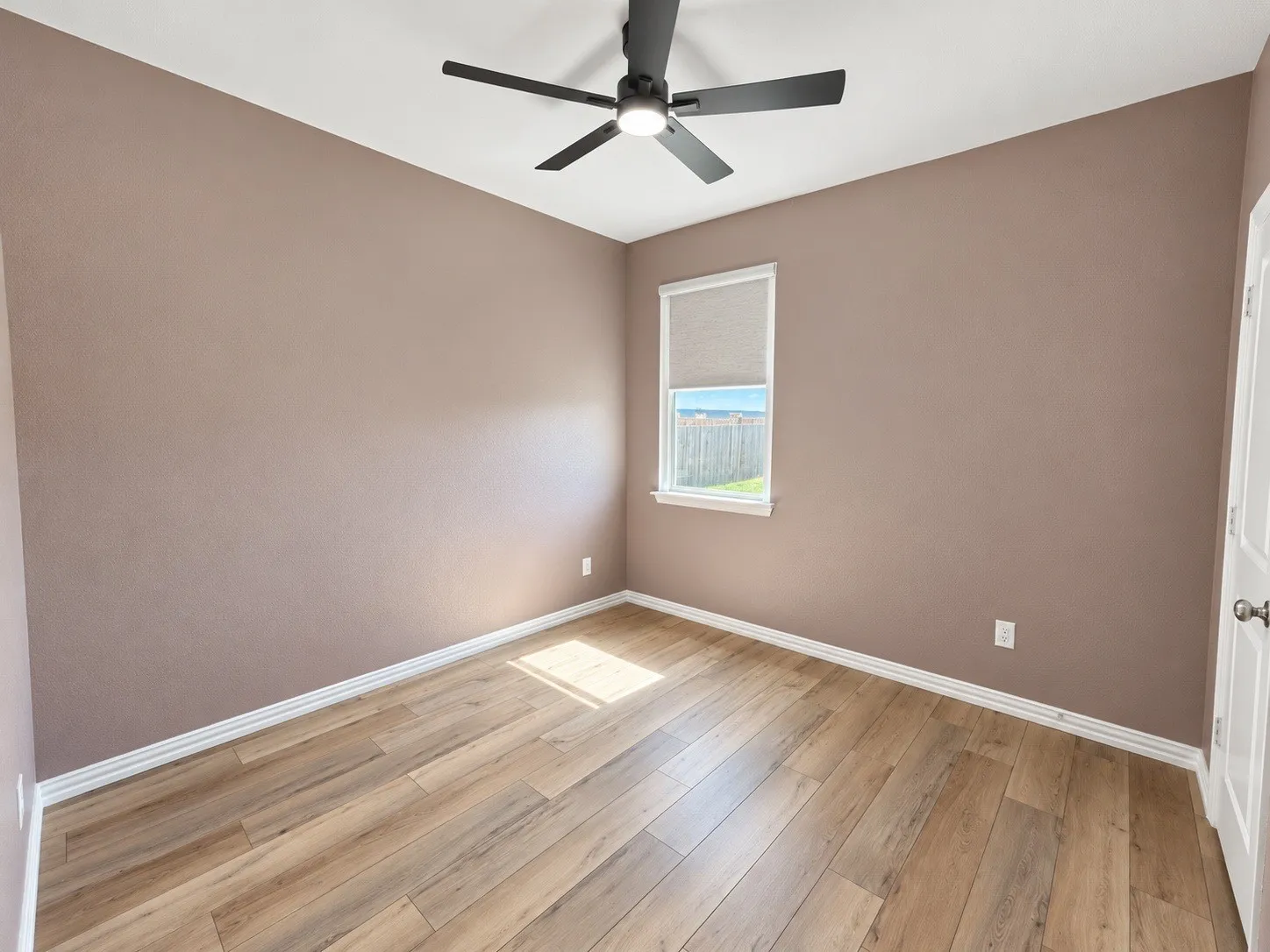 Neutral-toned walls, wood-finish flooring, ceiling fan, and a single window with a roller shade