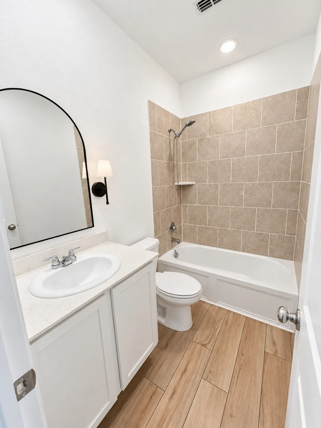 Wood-finish flooring, white vanity with integrated sink, chrome faucet, white bathtub with a tile surround, and a wall-mounted light fixture