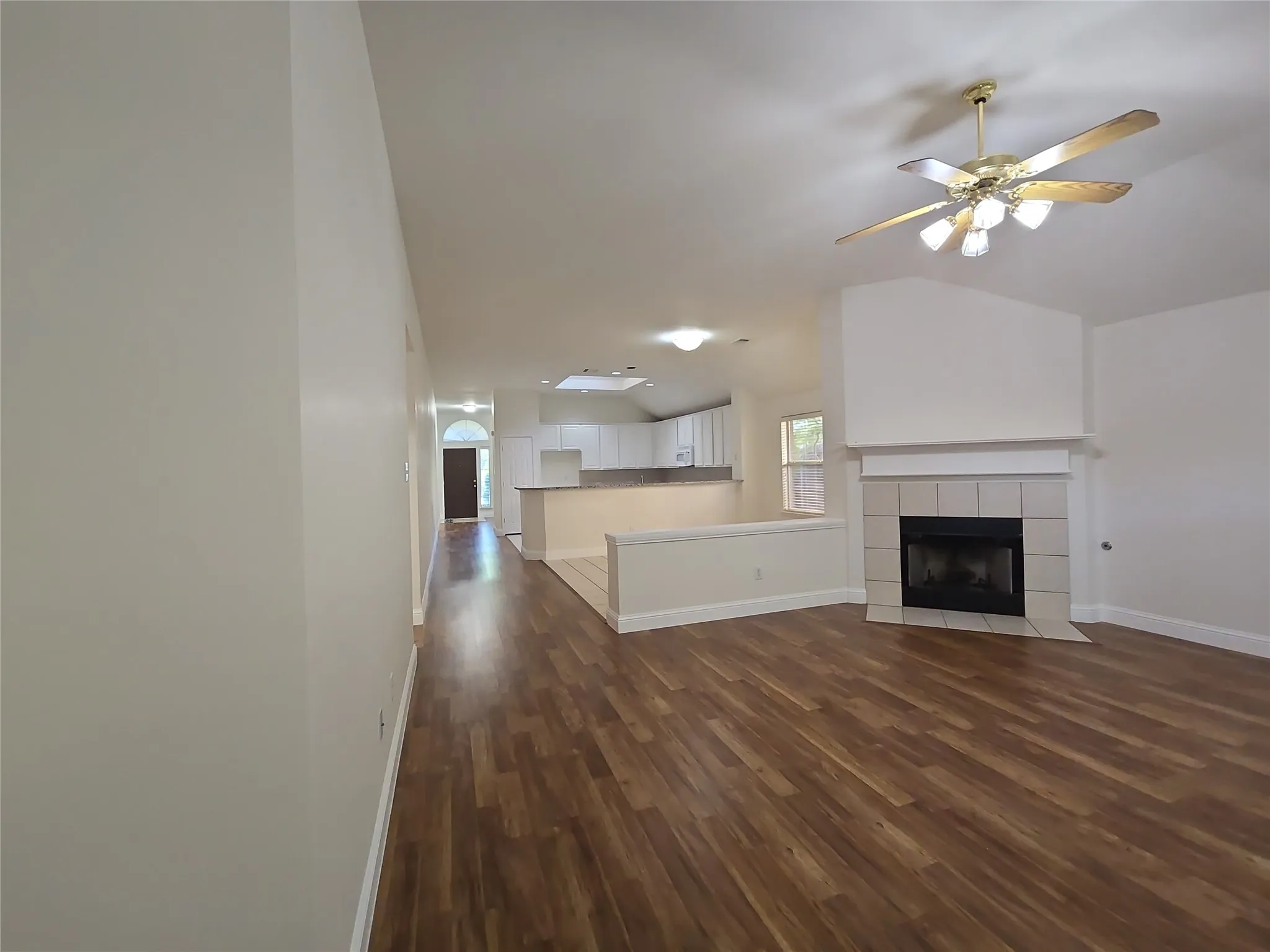 Vaulted ceiling with ceiling fan, fireplace with tiled surround, wood-finish flooring, and a kitchen with white cabinetry