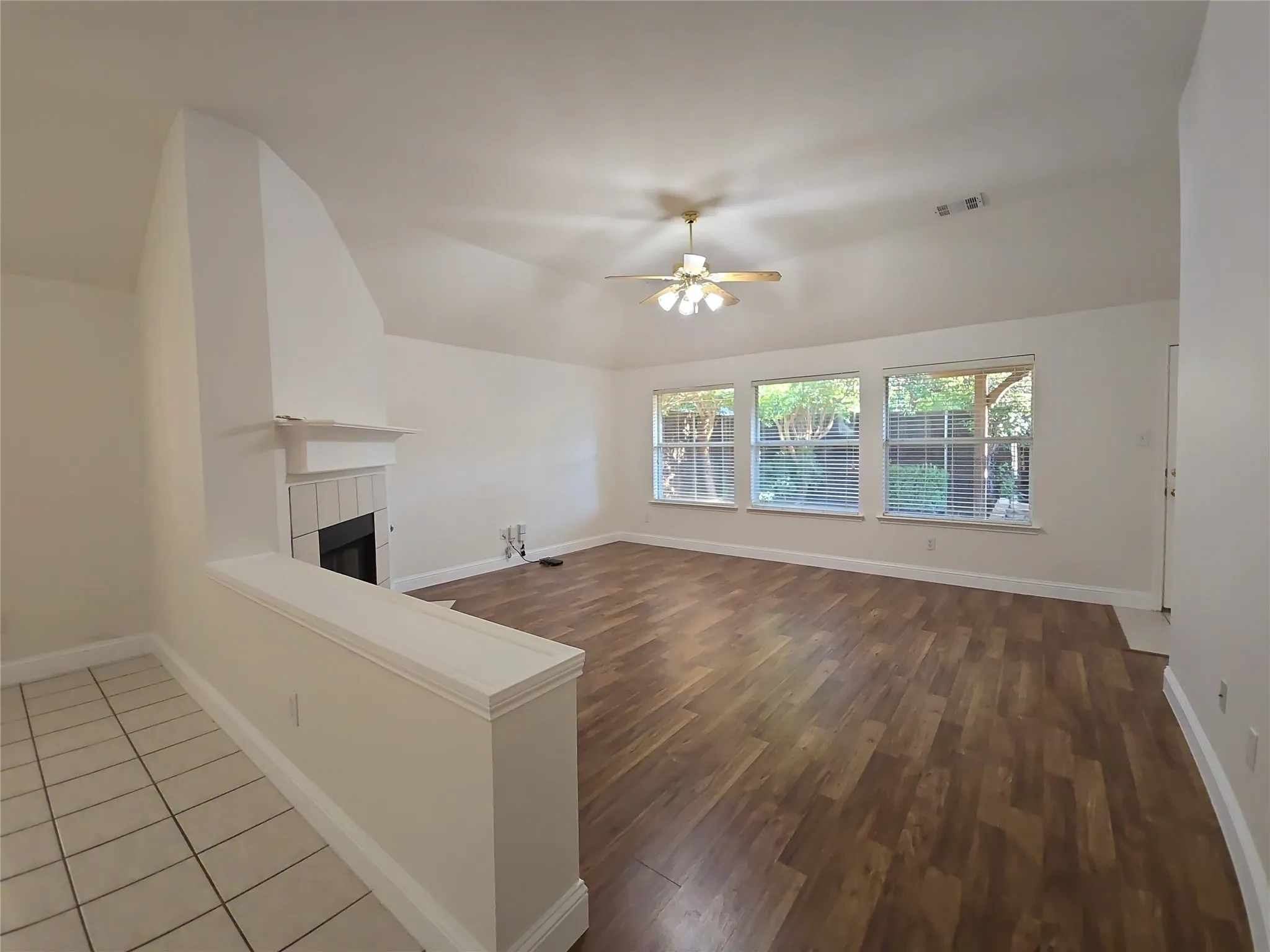 Open-concept living area featuring a vaulted ceiling with a ceiling fan, wood-finish flooring, a tile-surround fireplace, and three large windows