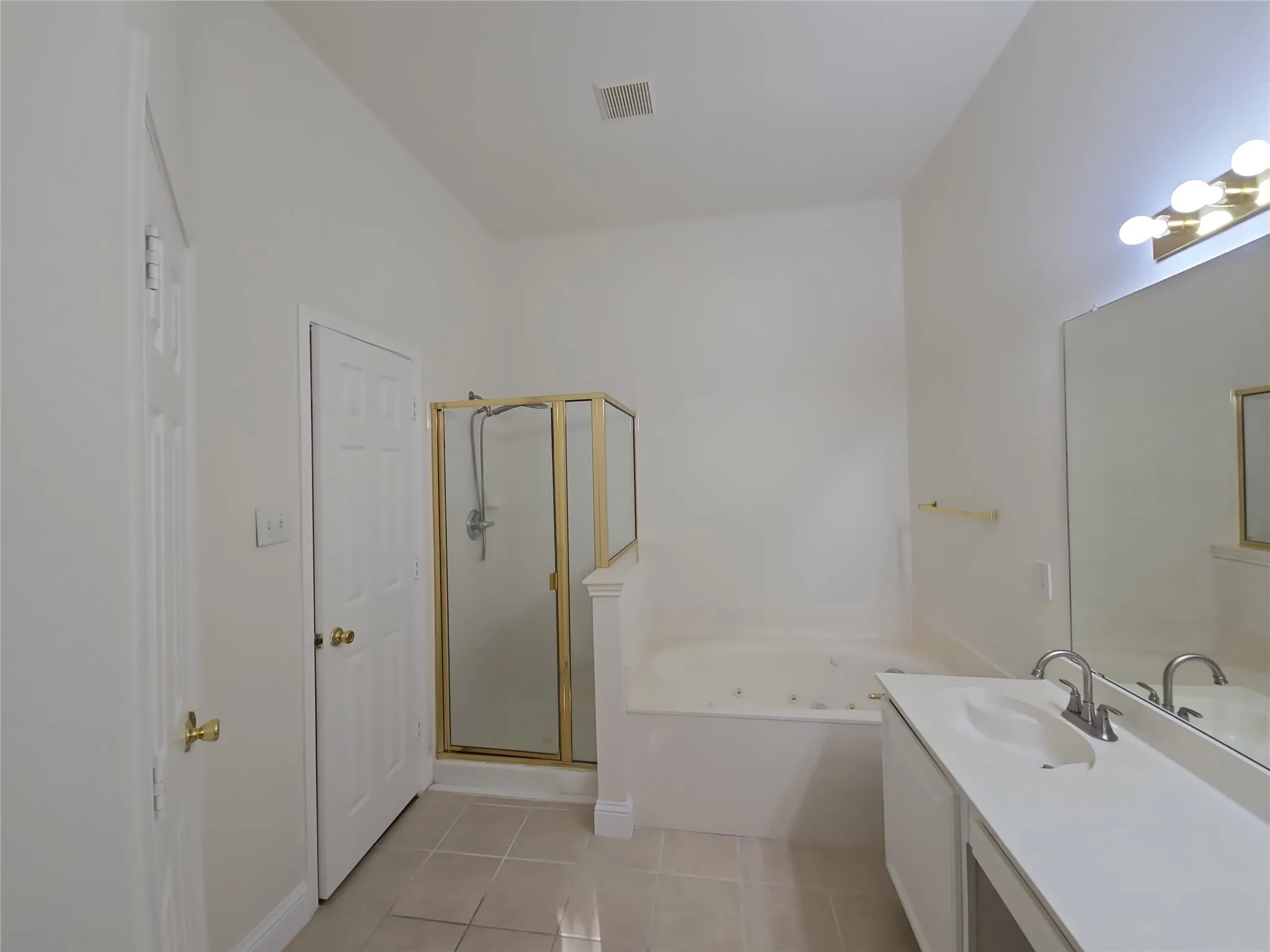 White paneled door, gold-framed shower enclosure, built-in bathtub, vanity with integrated sink, and large mirror with overhead lighting
