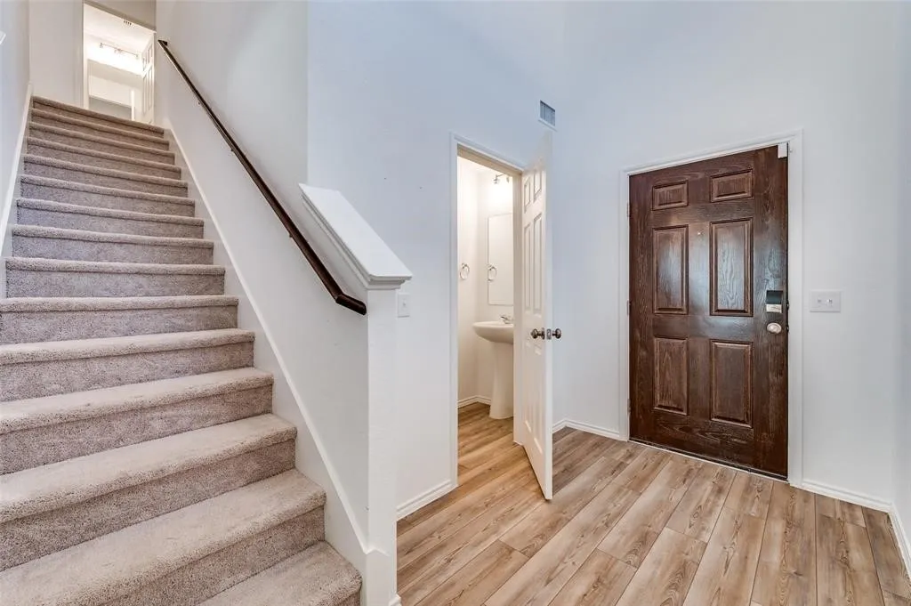Wood-finish flooring, dark wood front door, carpeted staircase with wood handrail, and a pedestal sink in the adjacent room