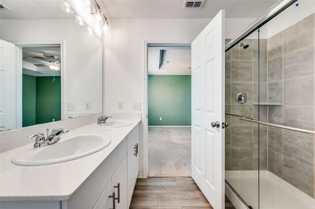 Dual vanity with white countertops, chrome fixtures, glass-enclosed shower with tile surround, and wood-finish flooring