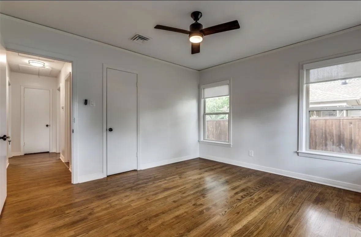 Rich wood-finish flooring, two windows with roller shades, a ceiling fan with integrated lighting, and a white interior door