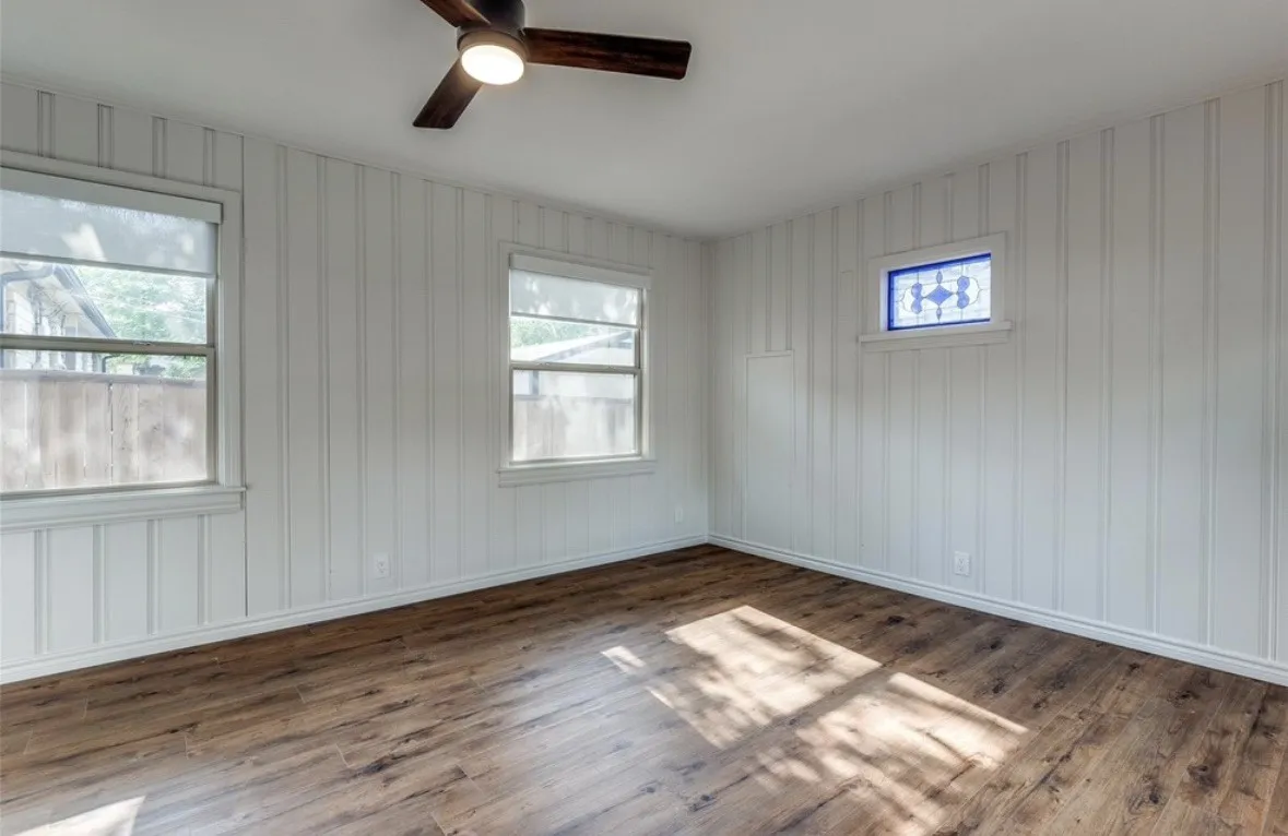 Wood-finish flooring, white vertical paneled walls, multiple windows, stained glass accent window, and a ceiling fan with integrated lighting