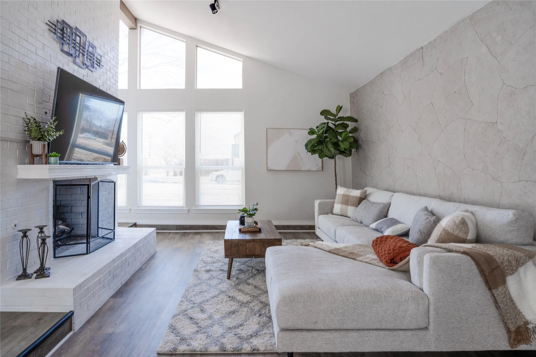 Vaulted ceiling with exposed beam, white brick fireplace with mantel, oversized windows with transoms, wood-finish flooring, and a textured accent wall