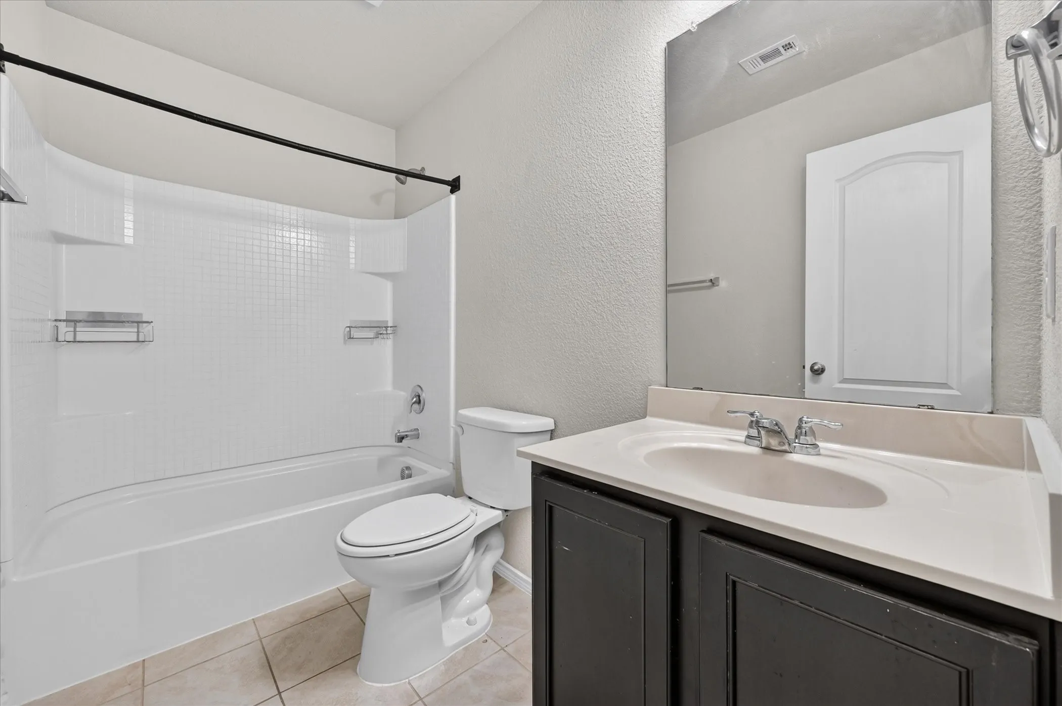 White bathtub with a black shower rod, white tile-patterned shower surround, dark wood vanity with white countertop, and integrated sink