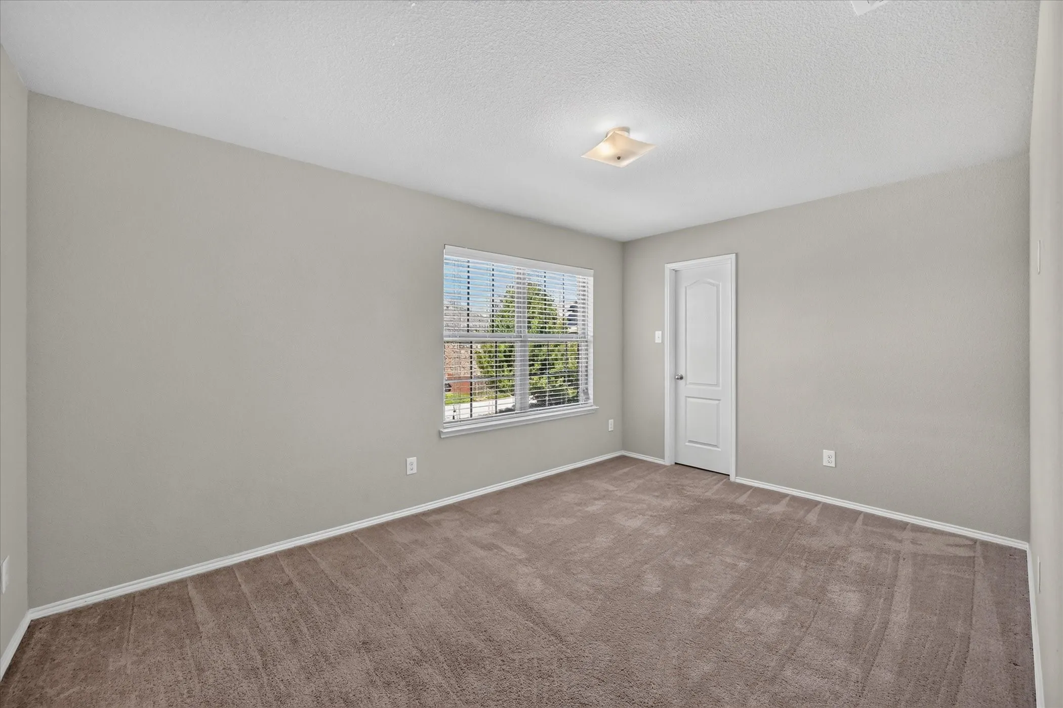 Neutral-toned walls, brown carpet, large window with blinds, white trim, and a flush-mount ceiling light
