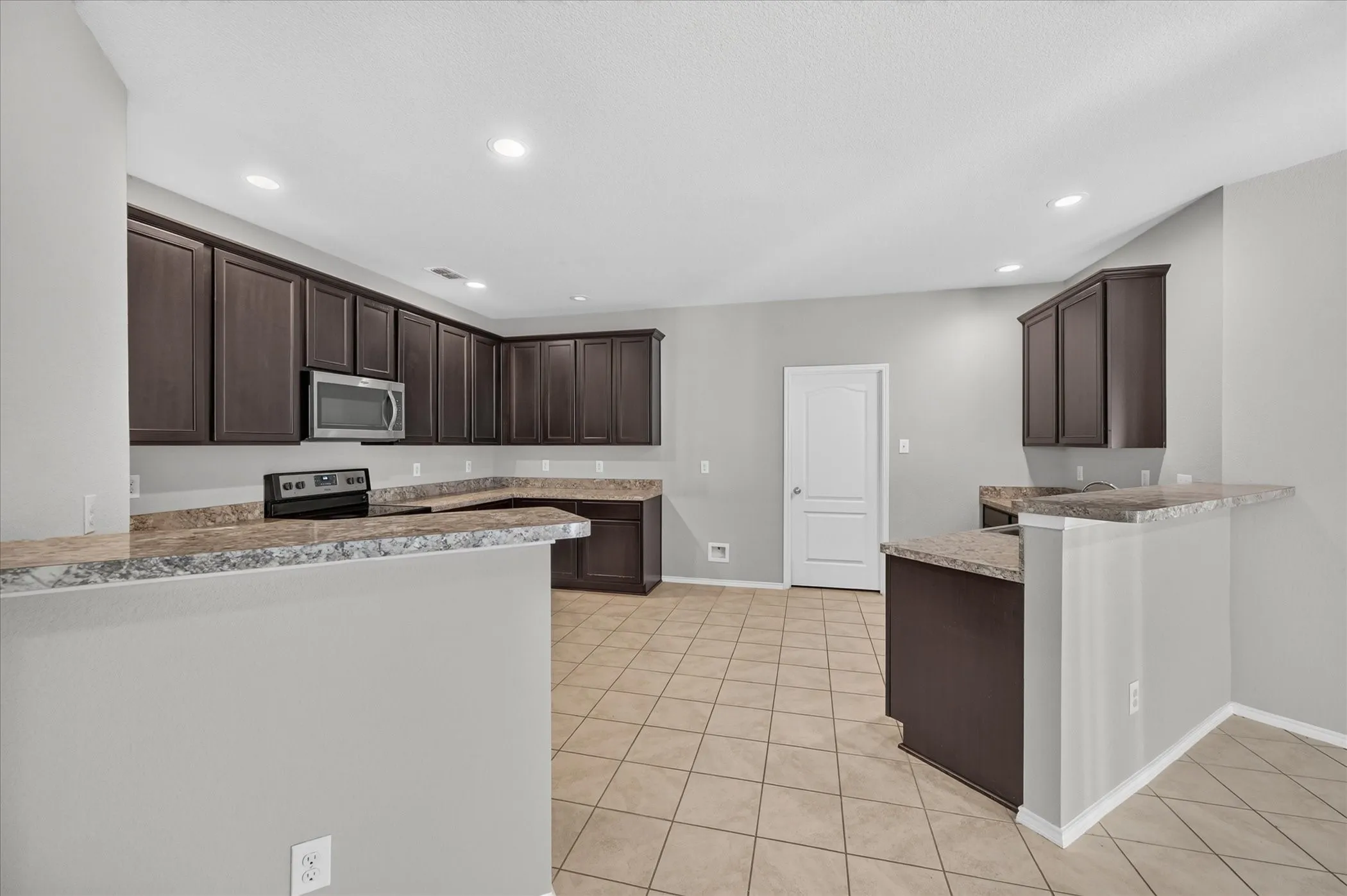 Kitchen featuring extensive dark wood cabinetry, light-toned tiled flooring, granite-style countertops, stainless steel microwave, and recessed lighting